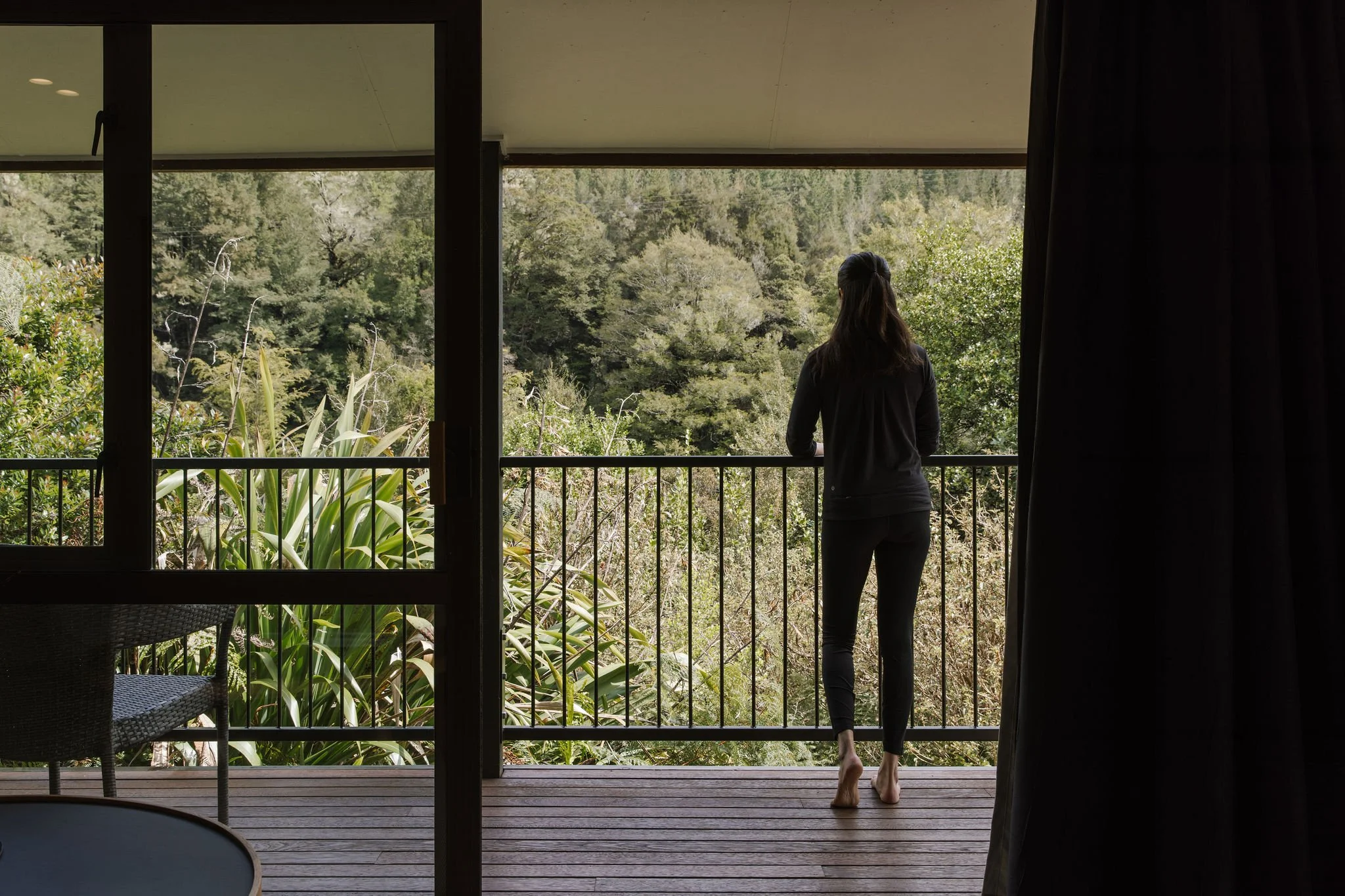 A woman standing barefoot on a balcony, looking at lush green foliage and trees in the distance.