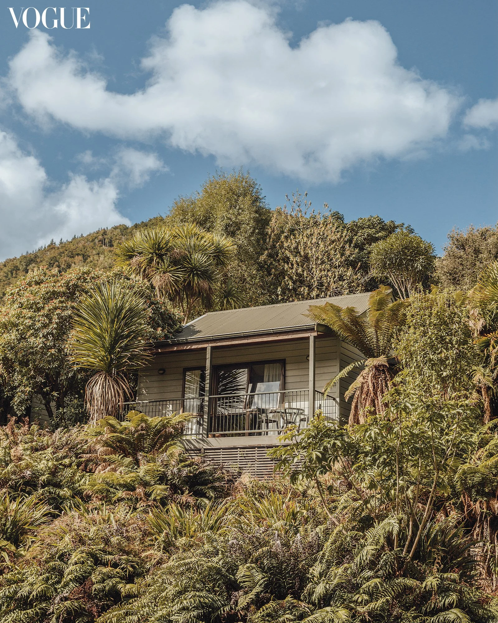 A house on a hillside surrounded by lush green trees and plants, with a blue sky and white clouds overhead.