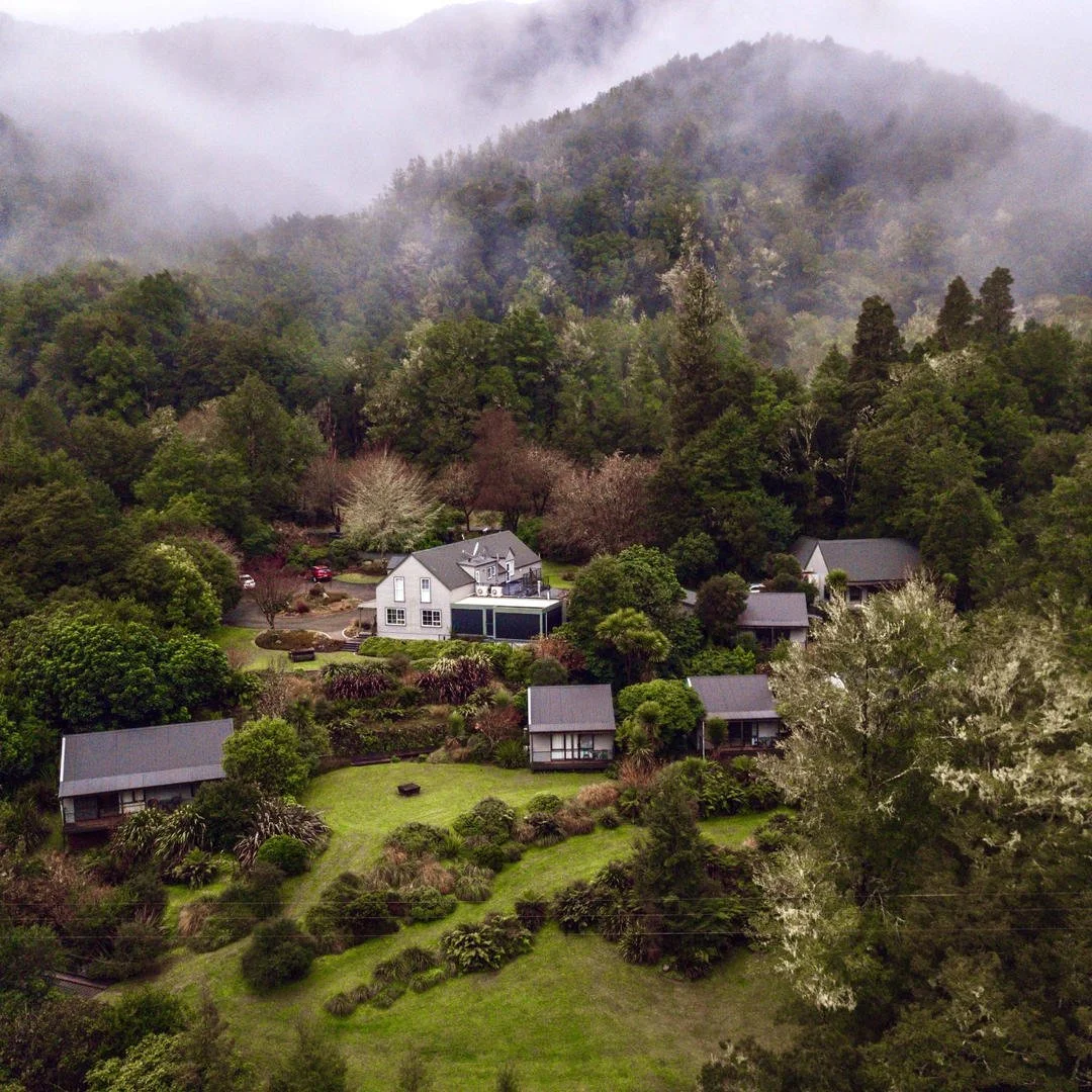 Aerial view of a rural hillside with multiple houses surrounded by lush green trees, under a cloudy sky with mist.