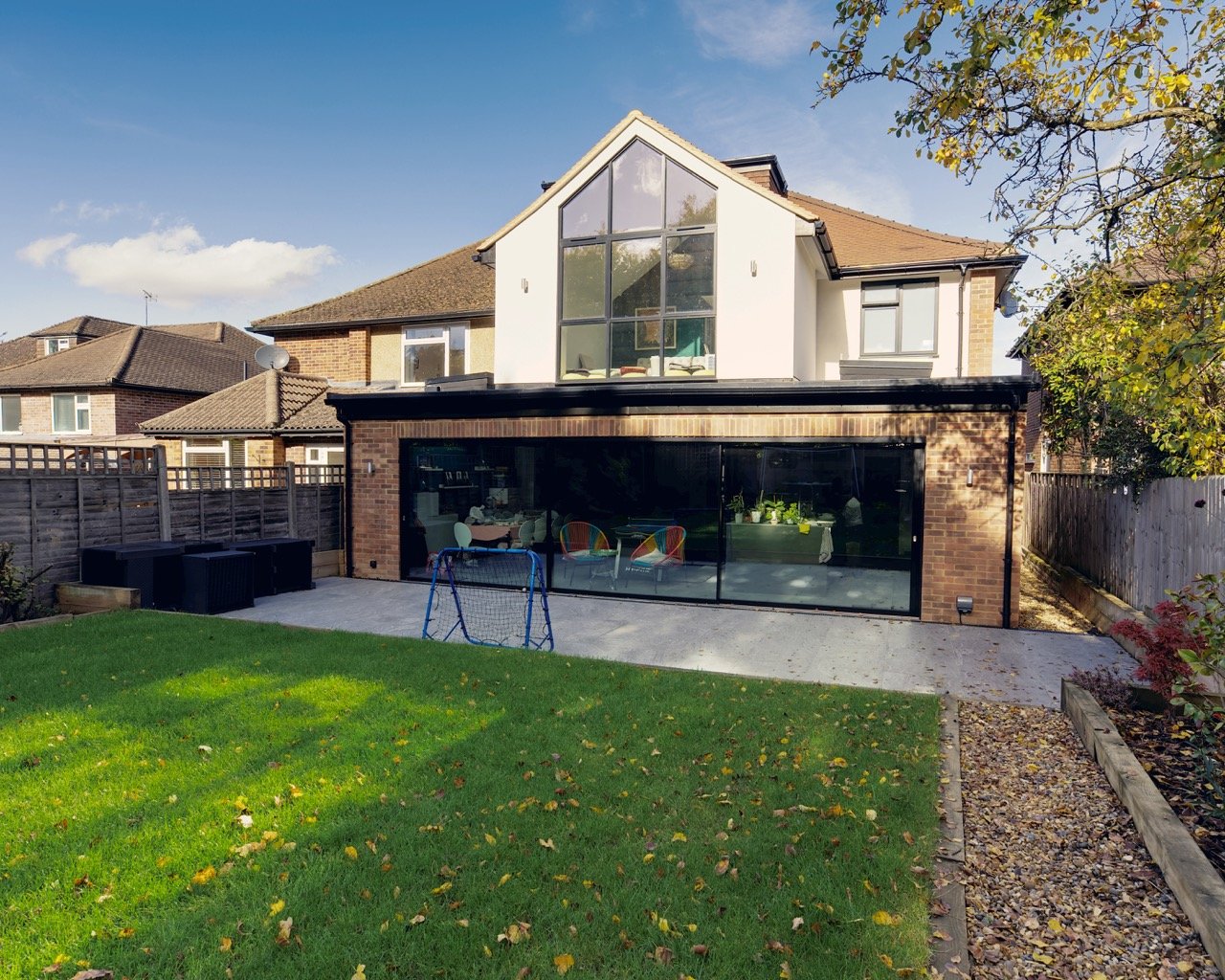 Modern two-story house with large glass windows, brick and white exterior, spacious backyard with green lawn, patio, and small soccer goal, surrounded by wooden fences and neighboring houses.