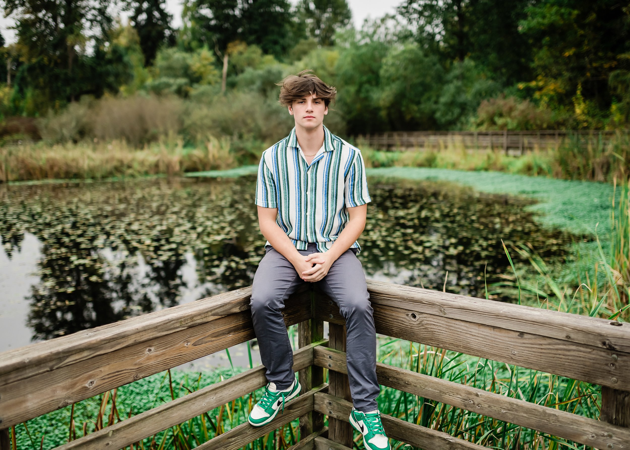  High school senior boy sitting on the railing in front of a lily pad filled lake. 