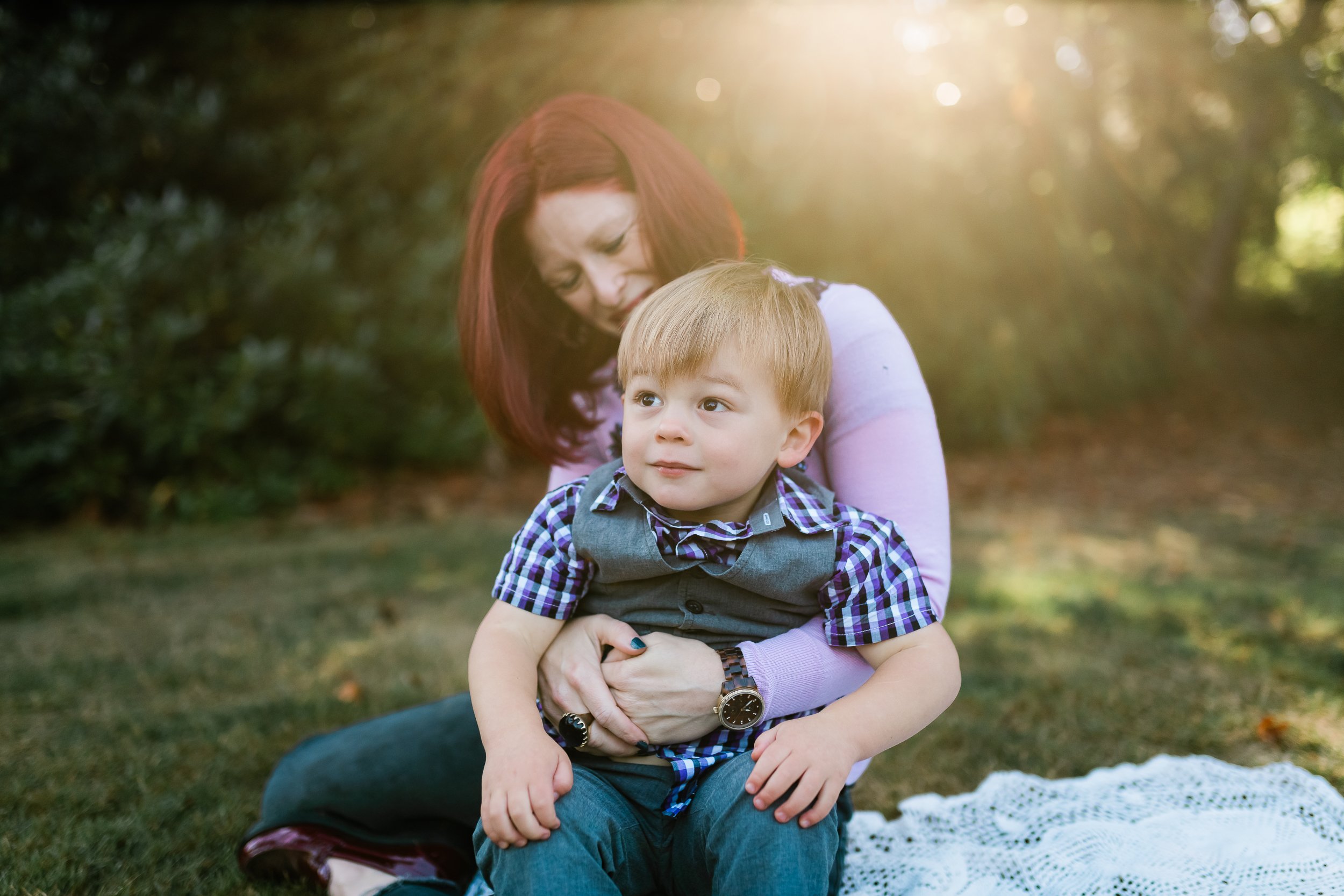  Mom holds toddler boy in her lap with sun behind her. 