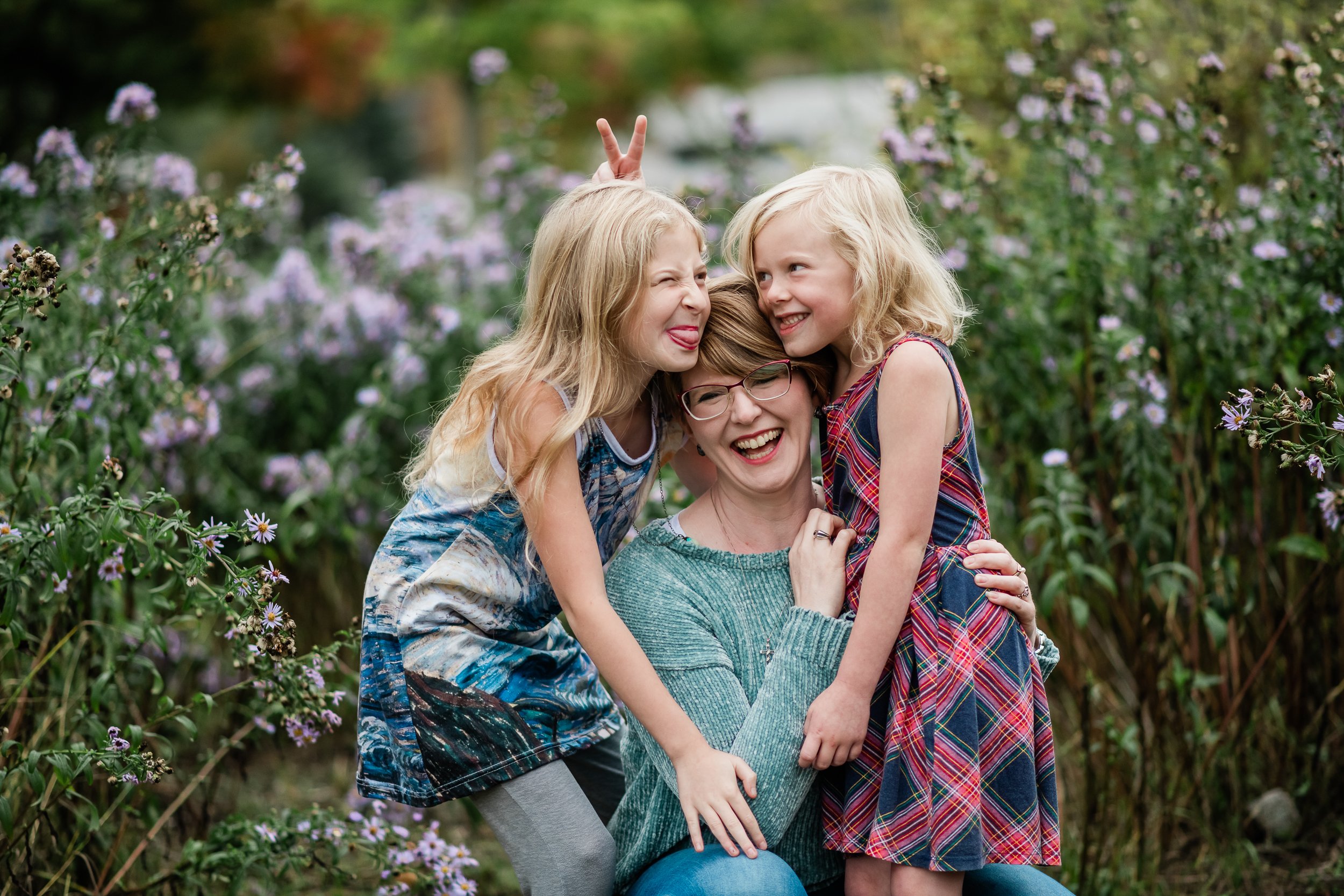  Laughing mother holds daughters in mismatched dresses while girls make faces at each other. 