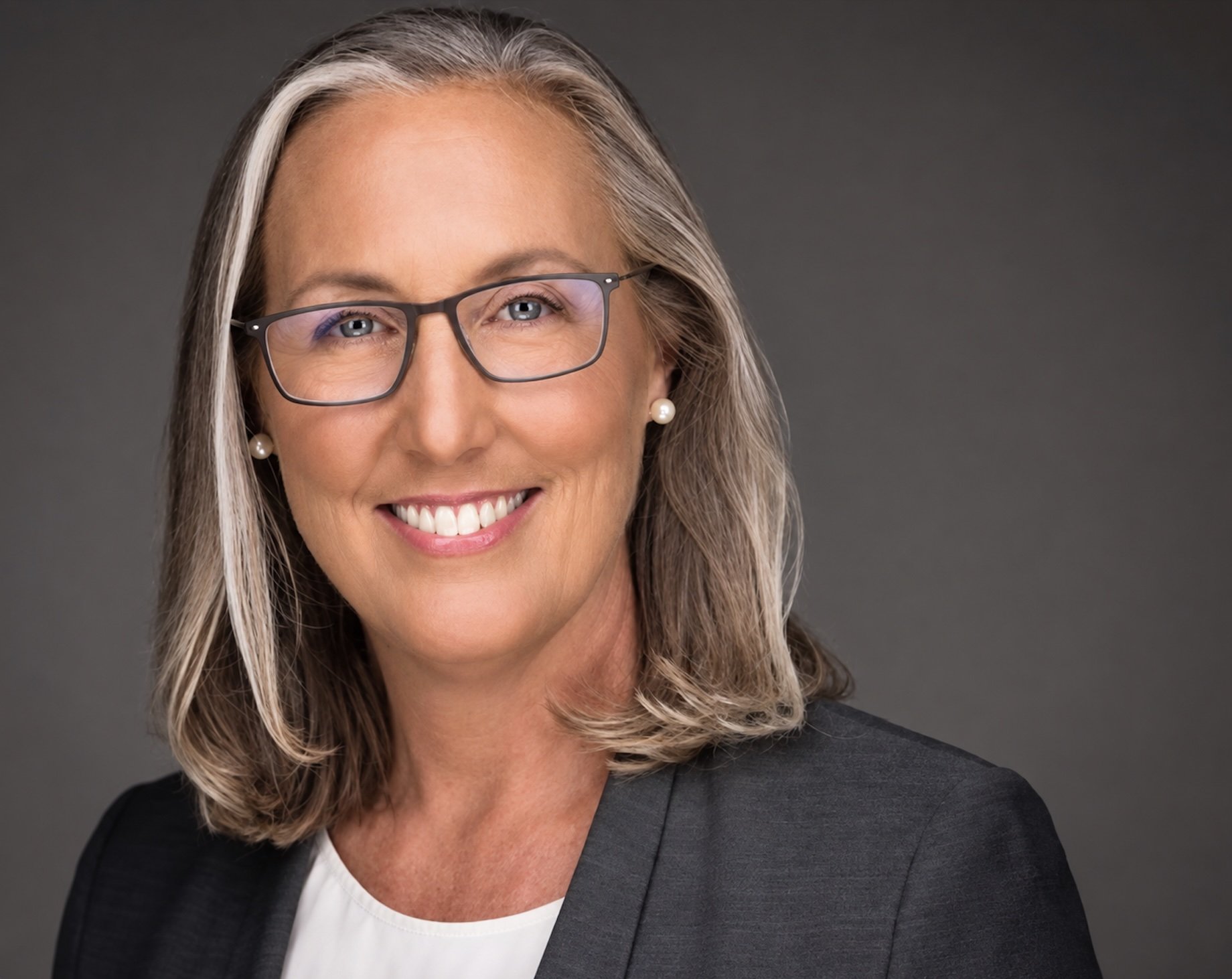 A woman in glasses and a dark grey suit smiles at the camera.