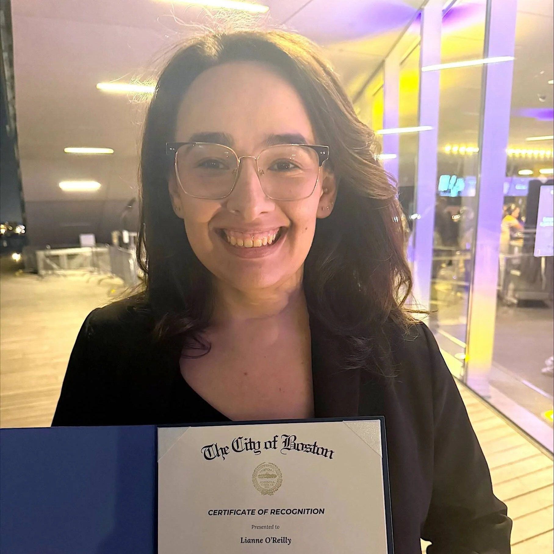 A woman with brown hair and glasses holds a certificate from the city of boston.