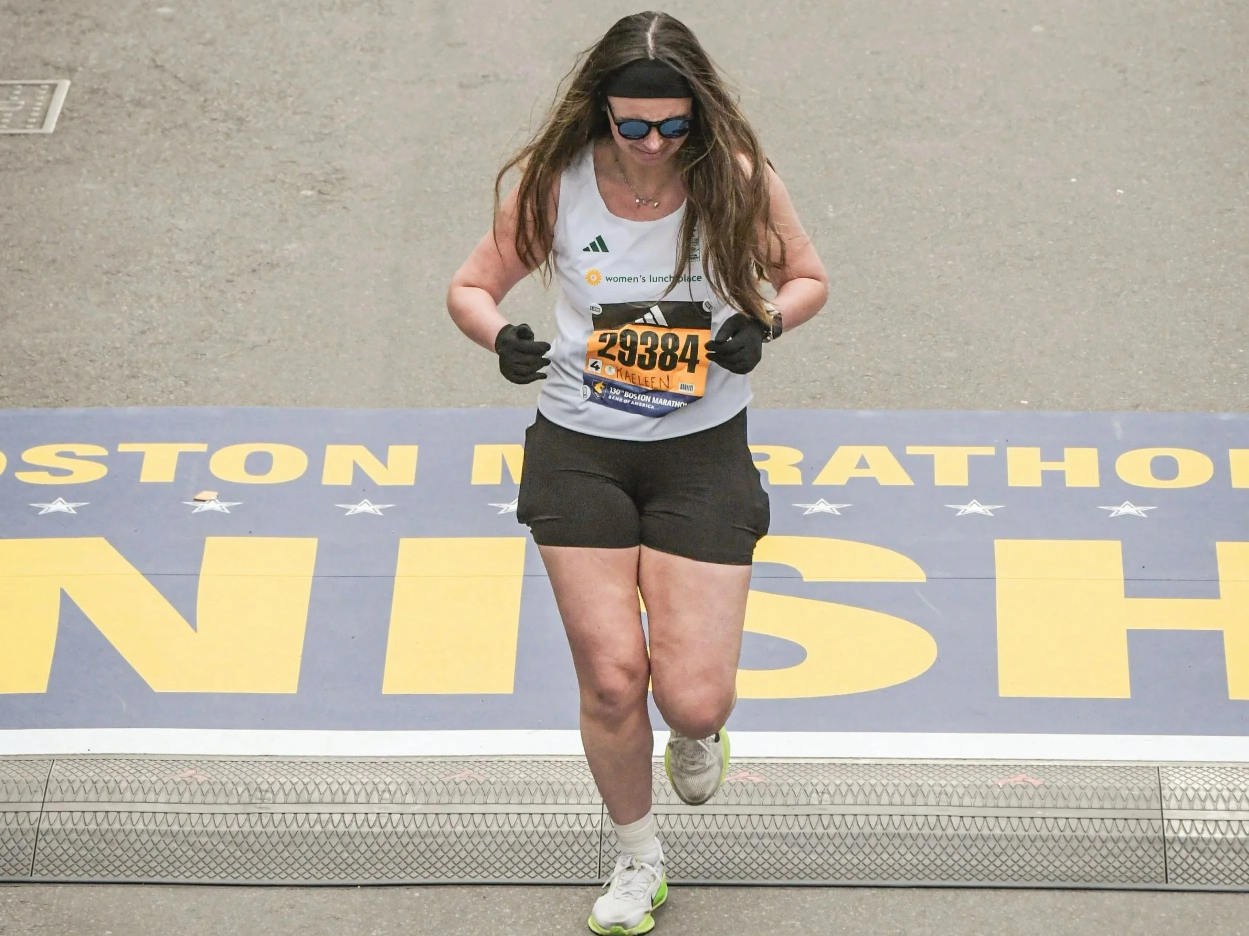 A woman in a white running jersey and black shorts crosses the finish line at the Boston Marathon.