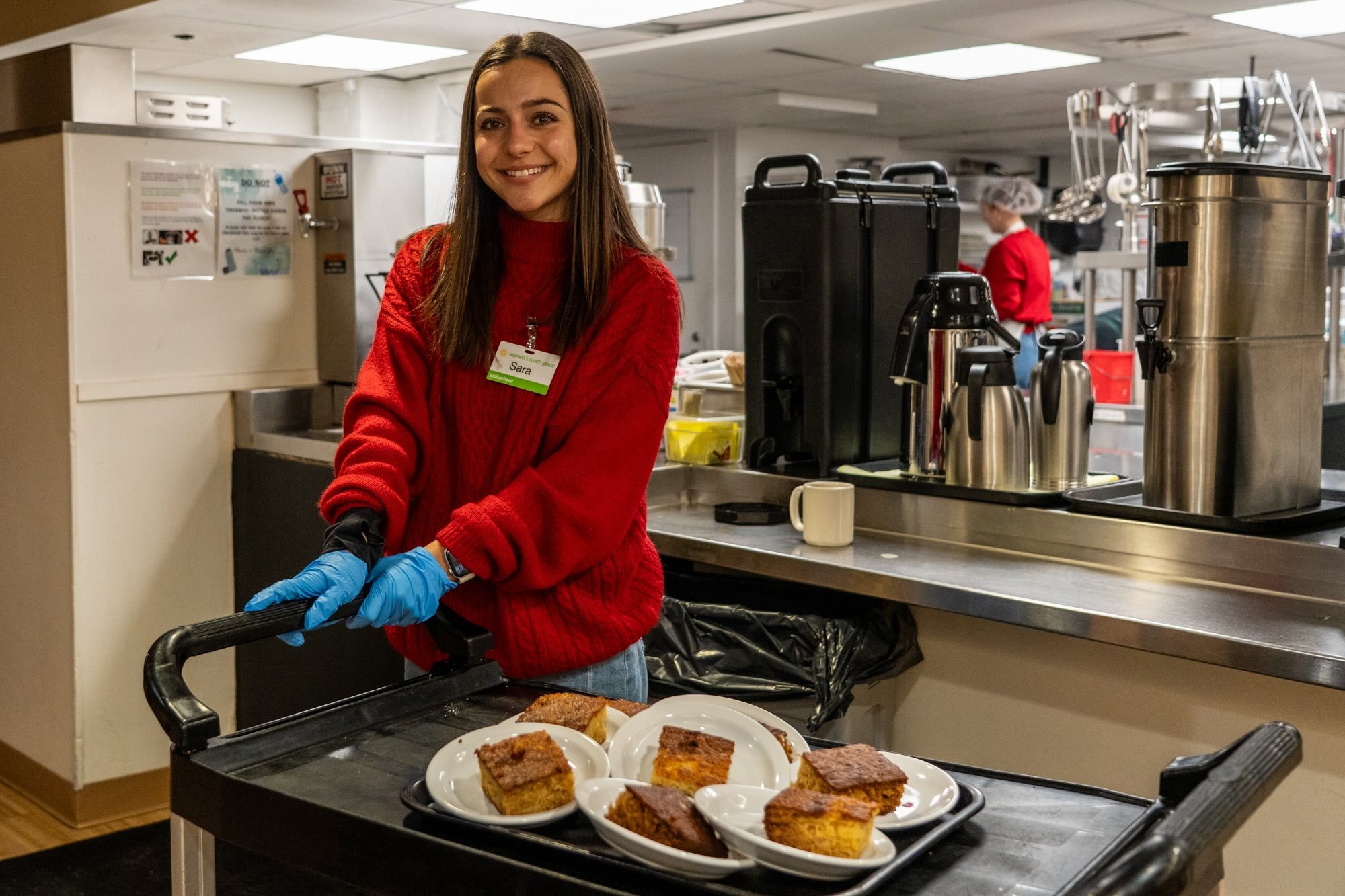 A volunteer wearing a red sweater standing in front of the kitchen with scratch-made desserts on a rolling cart.
