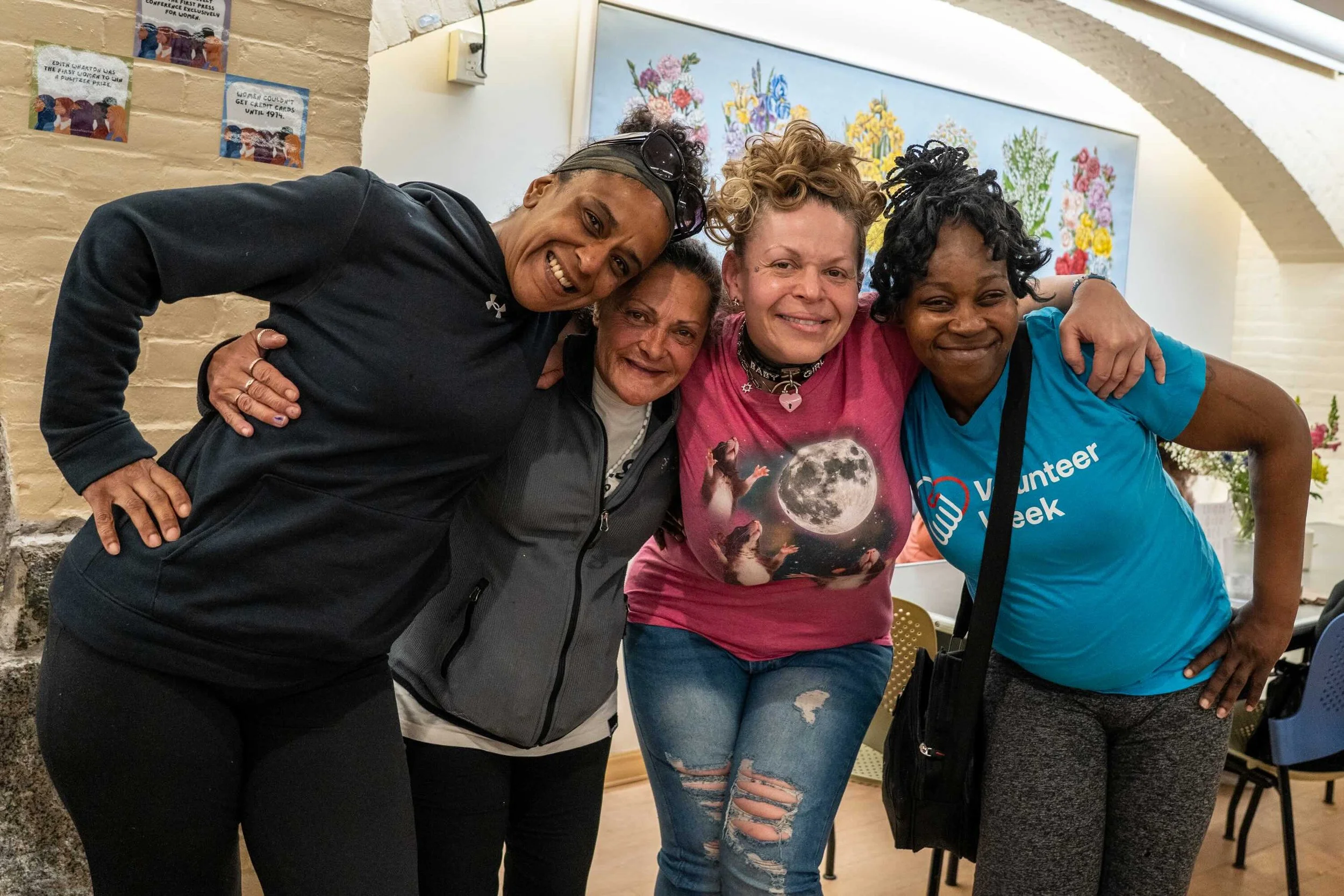 Four guests, arm in arm, smiling in the dining room of women's lunch place.