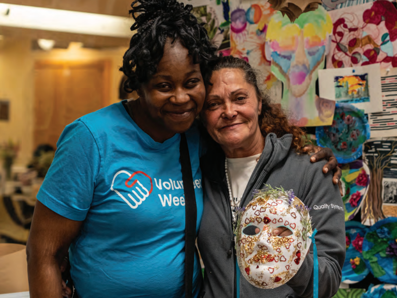 Two guests of Women's Lunch Place smiling. The guest on the right is holding a decorative mask that she has created in our Therapeutic Expressions program.