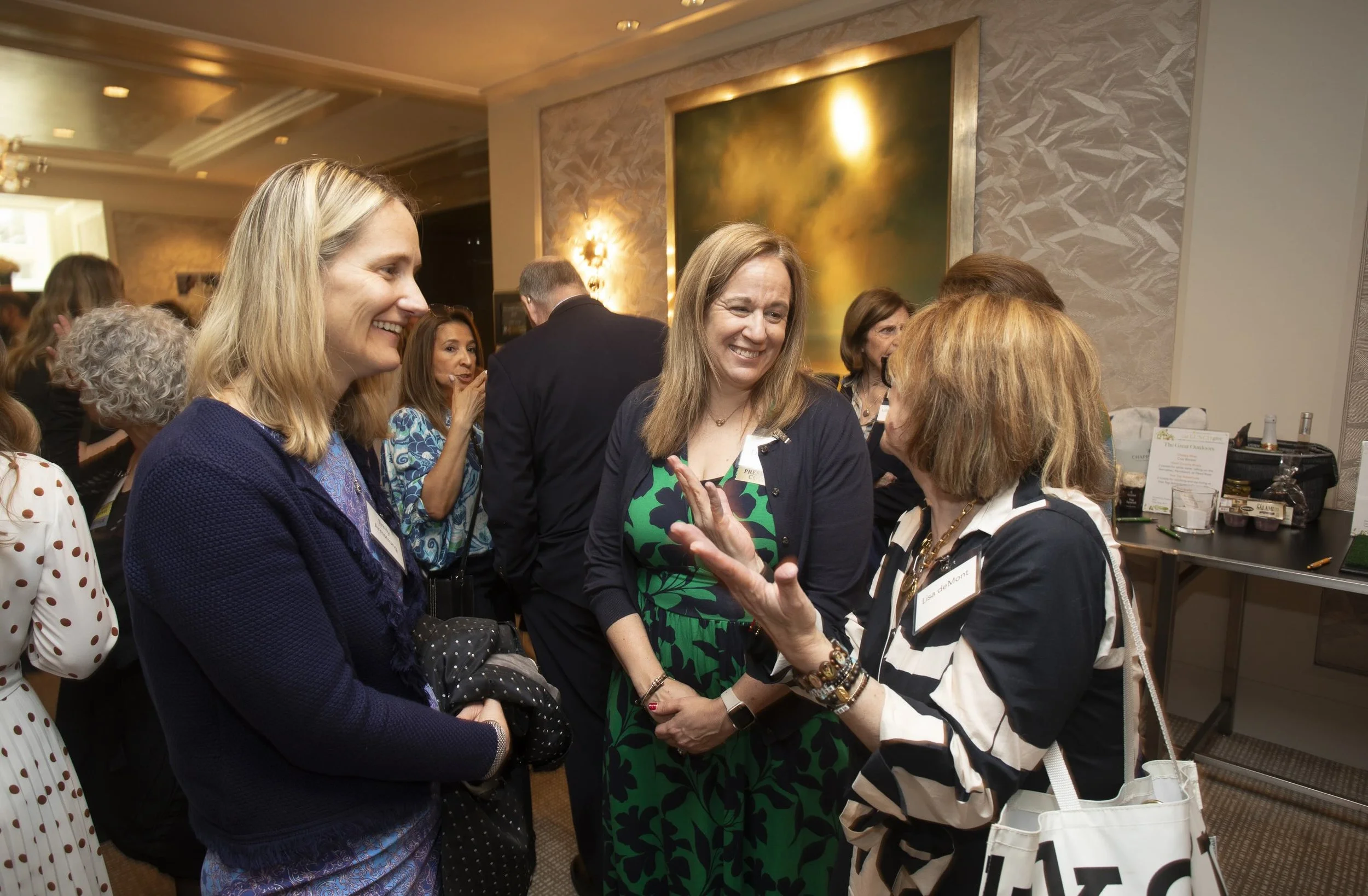 Three women smiling in conversation in a crowded room at last year's event.