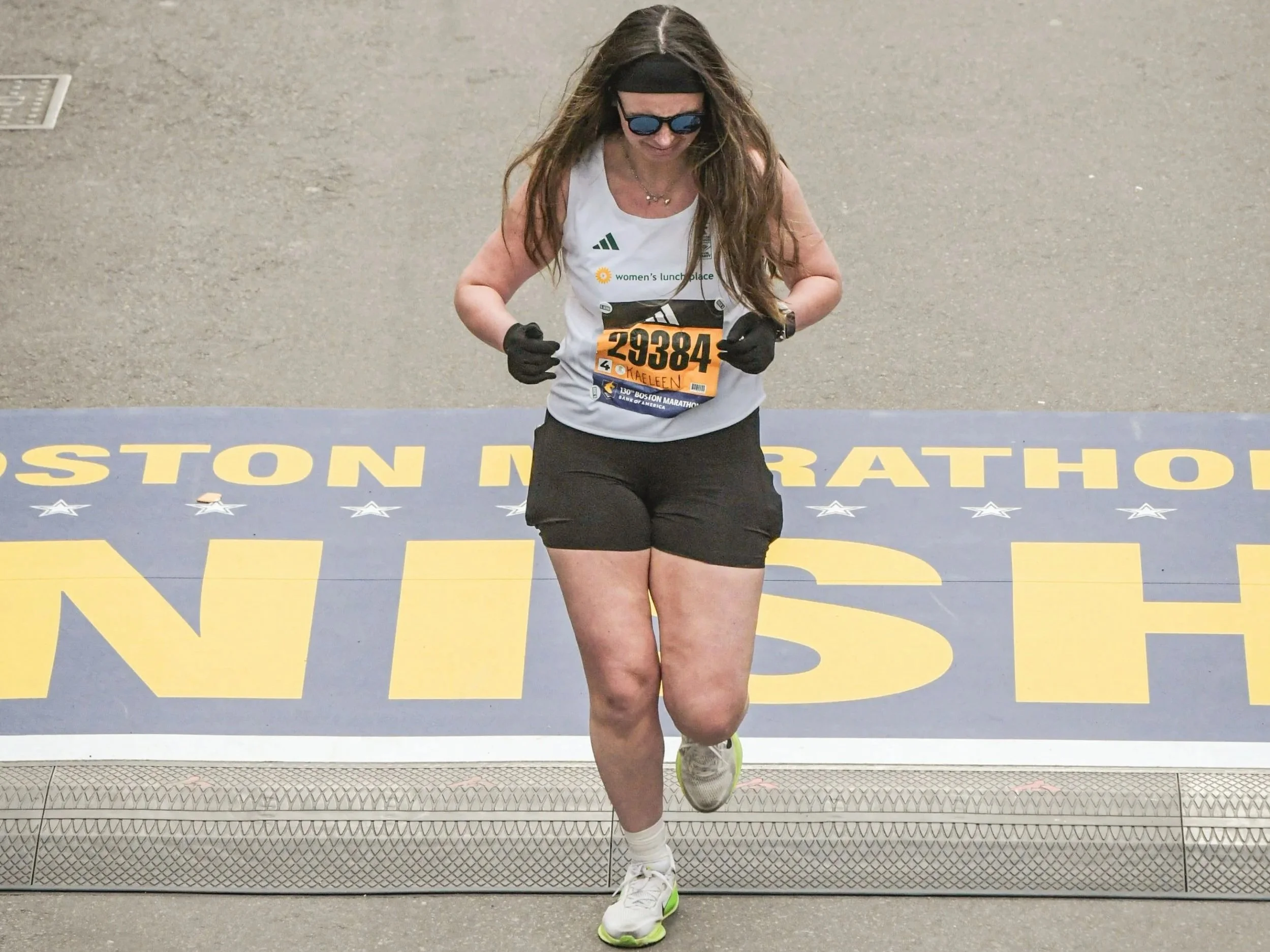 A woman in a white running jersey and black shorts crosses the finish line at the Boston Marathon.
