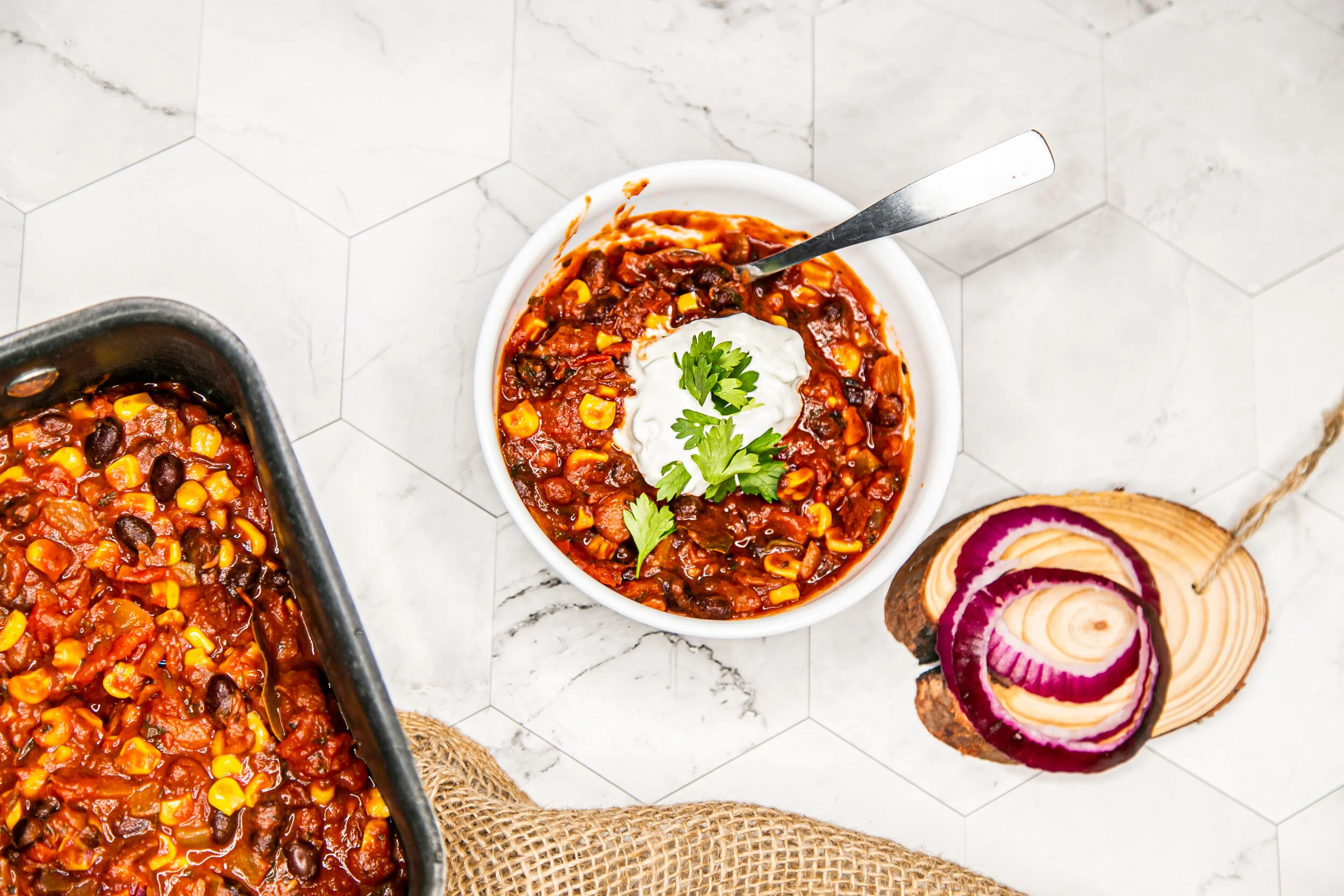 Bowl of chili topped with sour cream and cilantro, with a serving spoon on a white plate, a baking dish of chili, and a wooden cutting board with onion slices on a white marble hexagon tile surface.