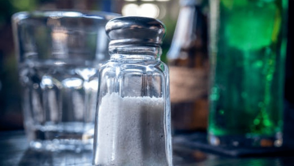Een glazen zoutvat gevuld met wit zout met op de achtergrond een glas en een groene fles