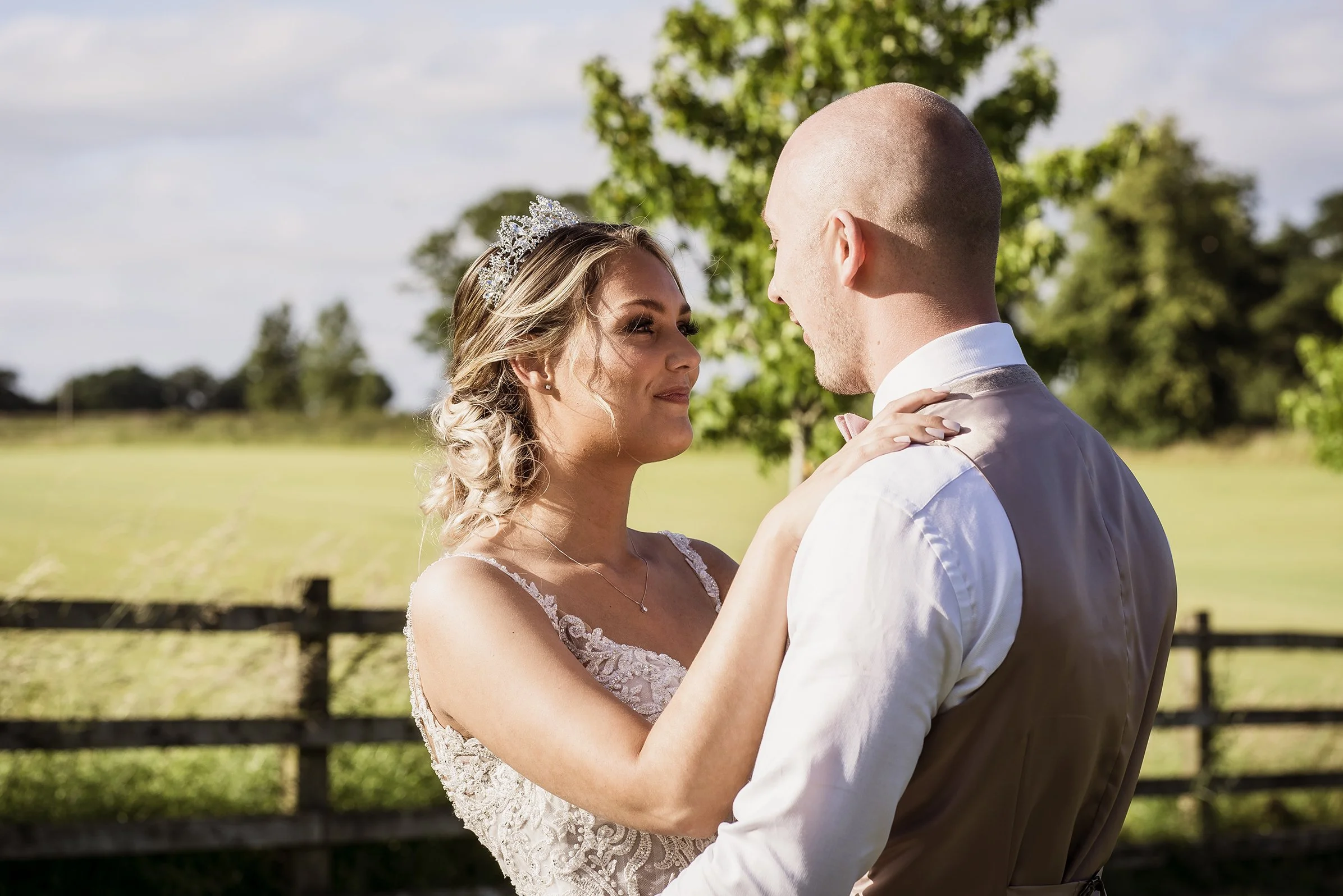 A bride and groom holding each other outdoors on their wedding day, with trees and a fence in the background at Sandhole Oak Barn