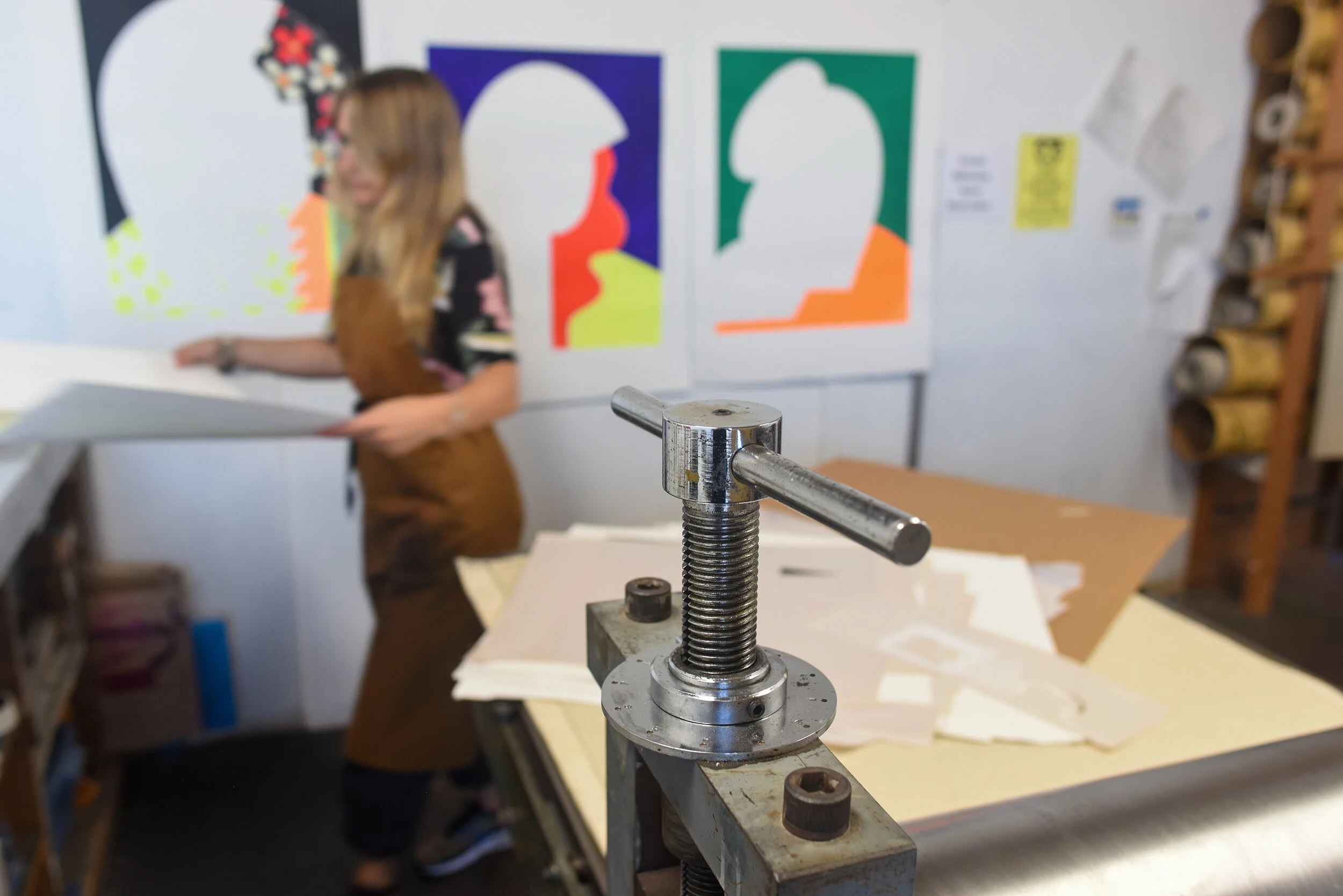 Close-up of a metal clamp screw on a worktable in a workshop, with a blurred woman in the background near colorful abstract posters on the wall.