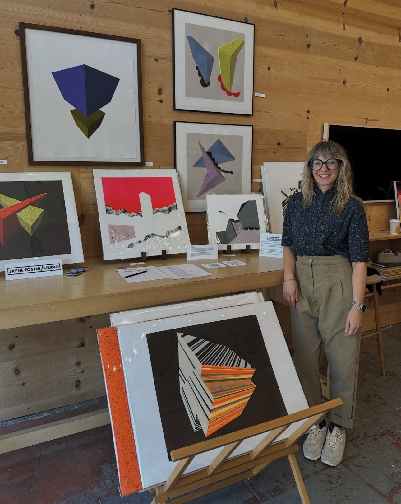 An art gallery with abstract geometric paintings displayed on a wooden wall and table. A woman with glasses, curly hair, a dark patterned shirt, beige pants, and white sneakers is smiling at the camera.