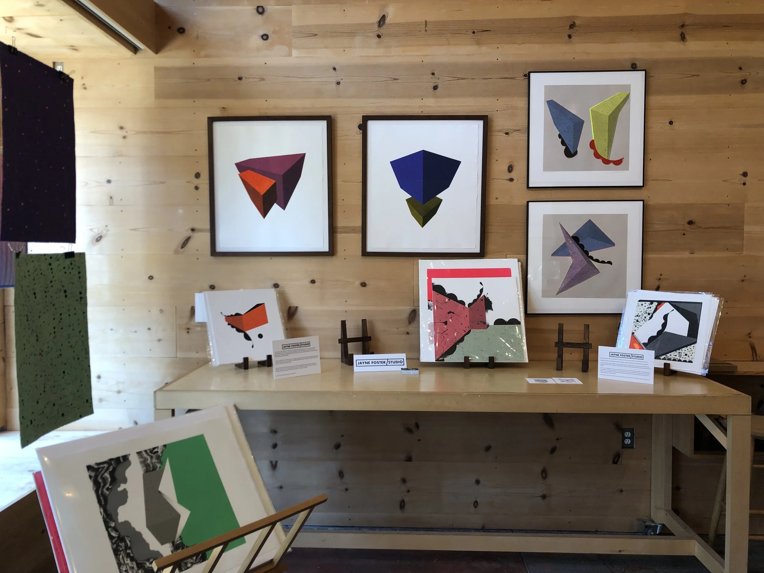 Art exhibit featuring colorful geometric abstract artwork displayed on a wooden wall and tables.