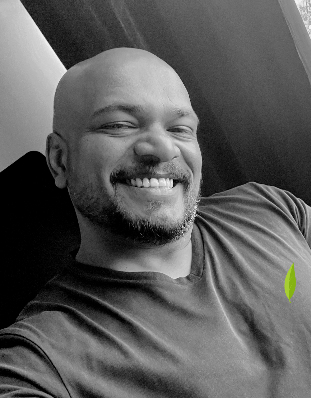 A smiling bald man with a beard, wearing a dark t-shirt, reclining and taking a selfie indoors.