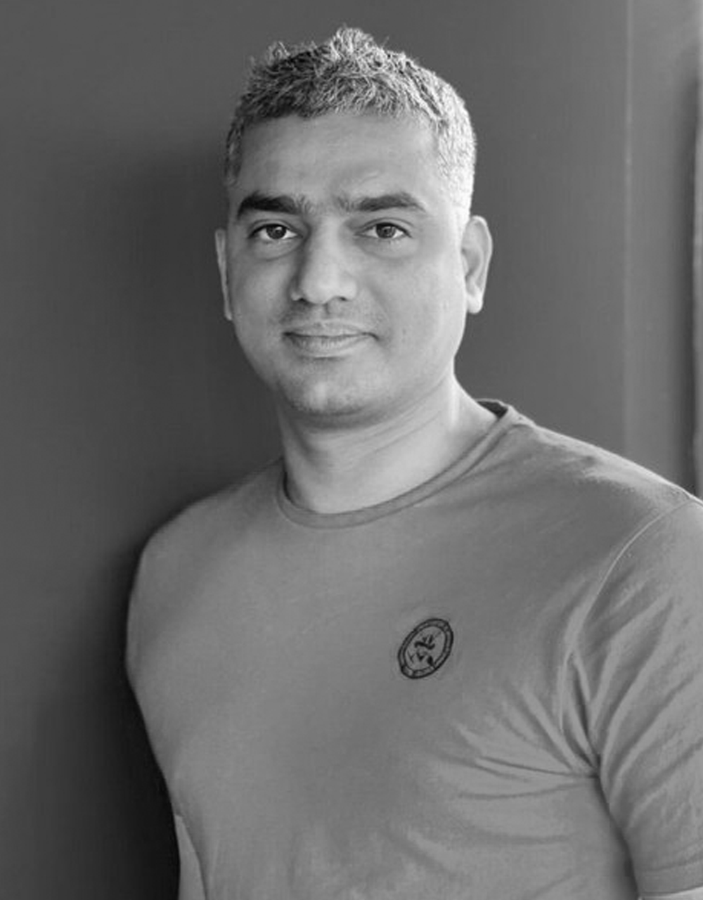 Black and white portrait of a young man with short, light-colored hair, wearing a t-shirt with a small emblem on the chest, standing against a plain wall.