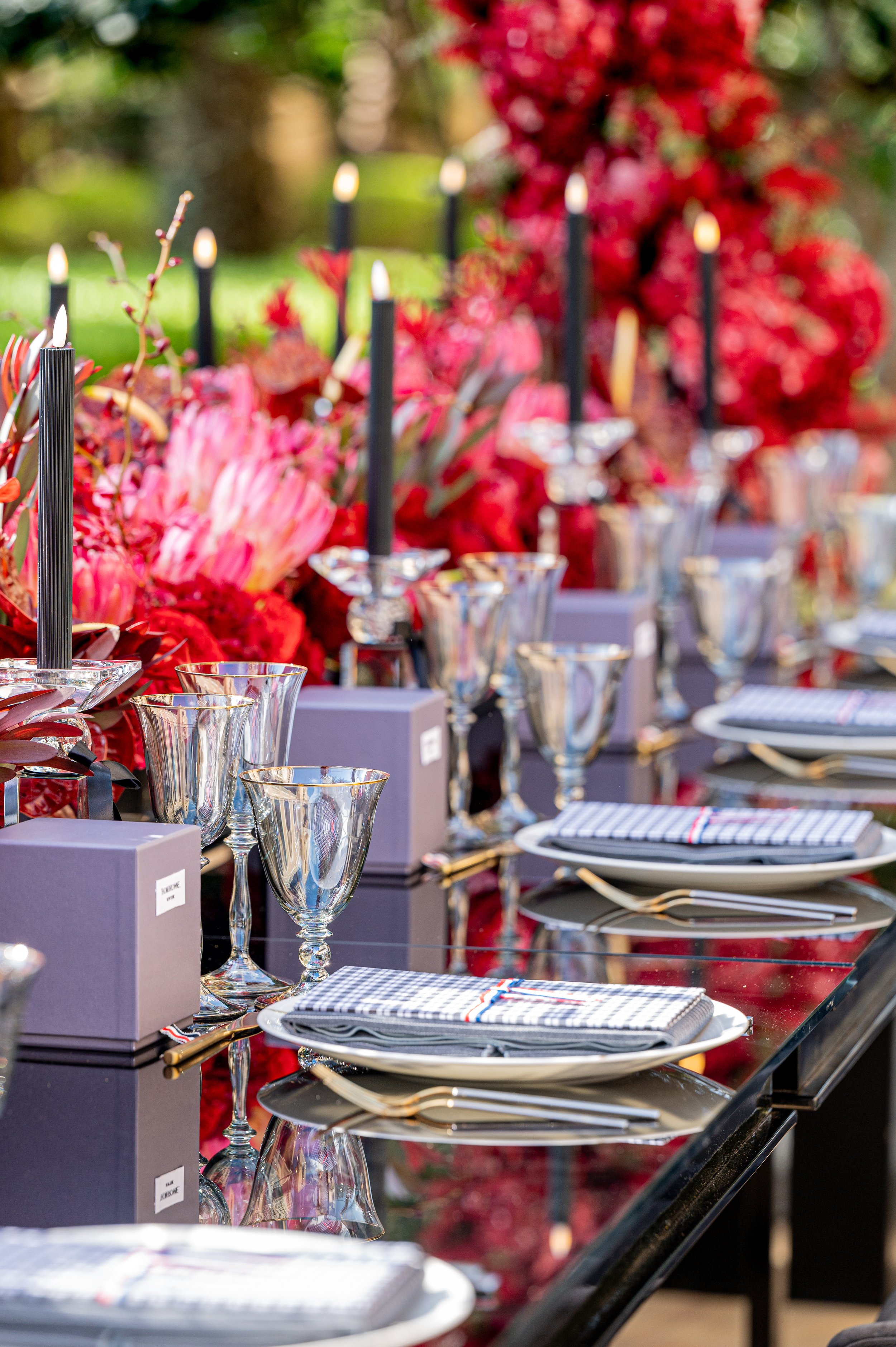 An elegant outdoor table setting with wine glasses, plates, silverware, and linen napkins, decorated with colorful flowers and black candles.