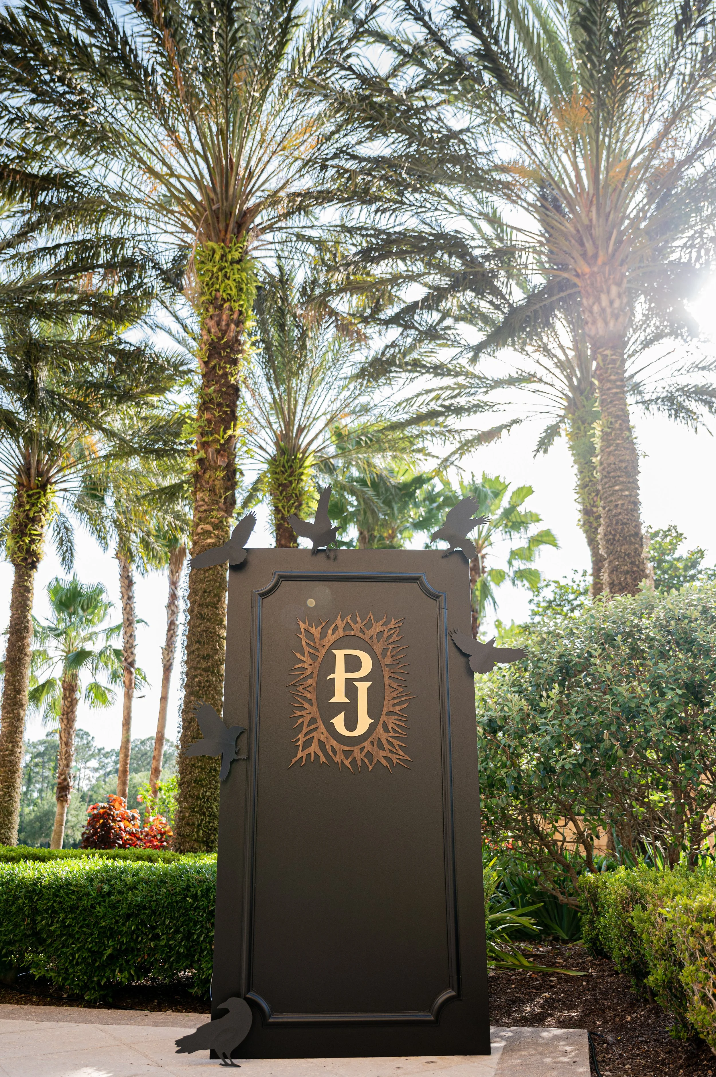 A black sign with the initials 'PJ' surrounded by bird silhouettes, set among palm trees and lush greenery in bright sunlight.
