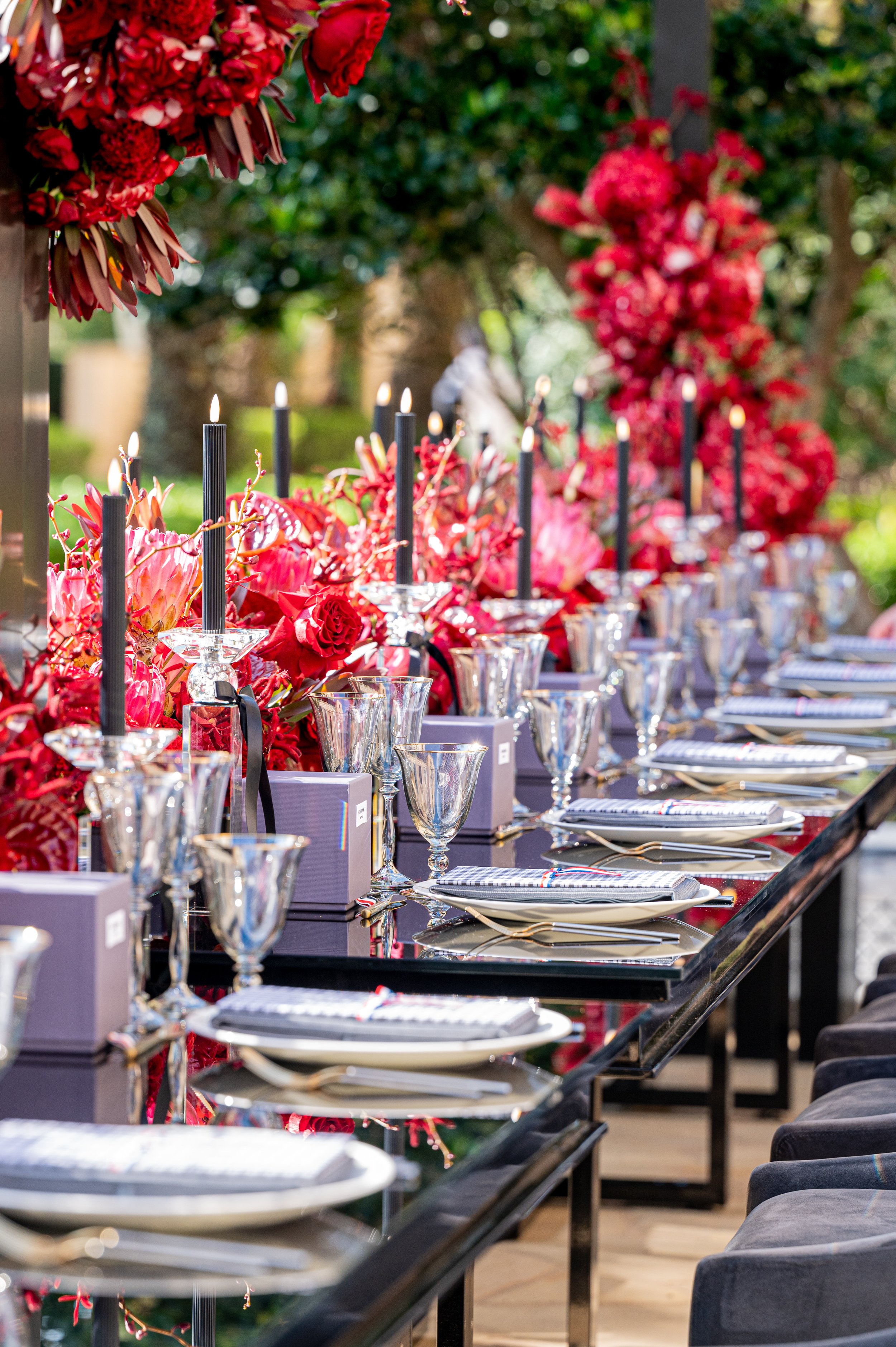 Elegant outdoor dining table decorated with red flowers, black candles, crystal glasses, and neatly arranged plates and utensils.