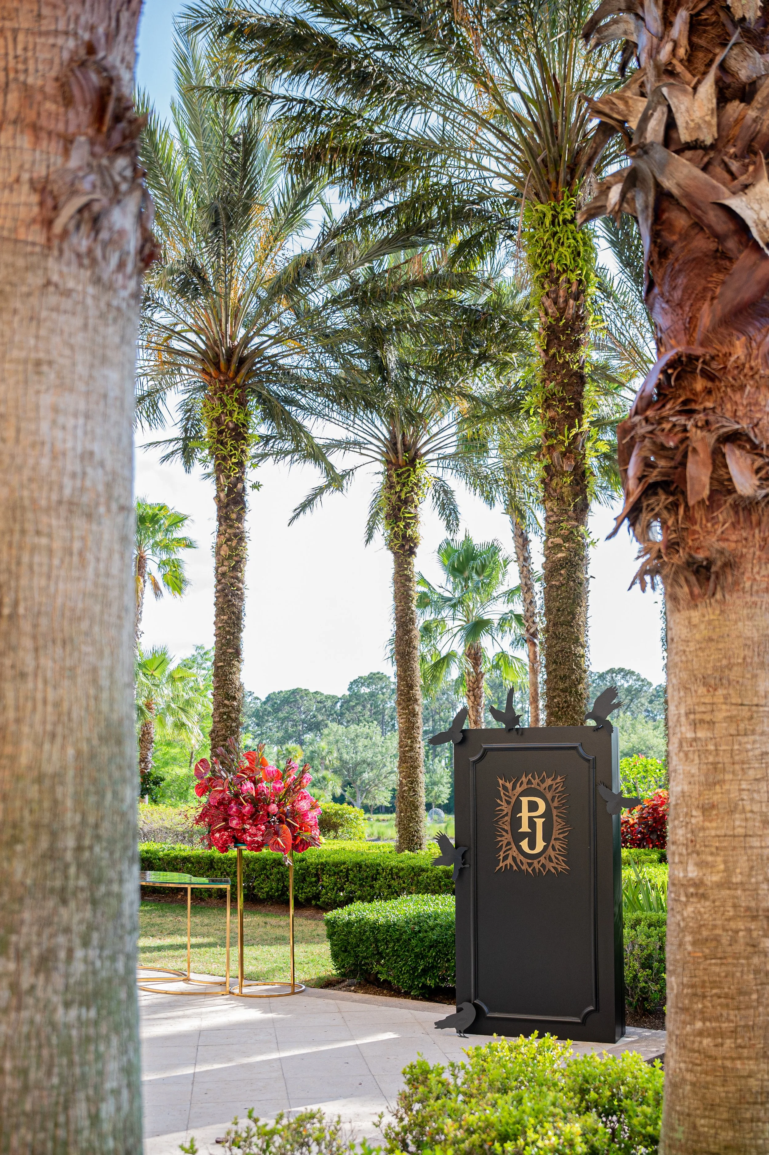 A lush garden with tall palm trees, trimmed bushes, and a black sign with gold lettering that reads 'PJ', decorated with black bird silhouettes. Pink flowers are visible near the sign, and the scene is sunny with a clear sky.