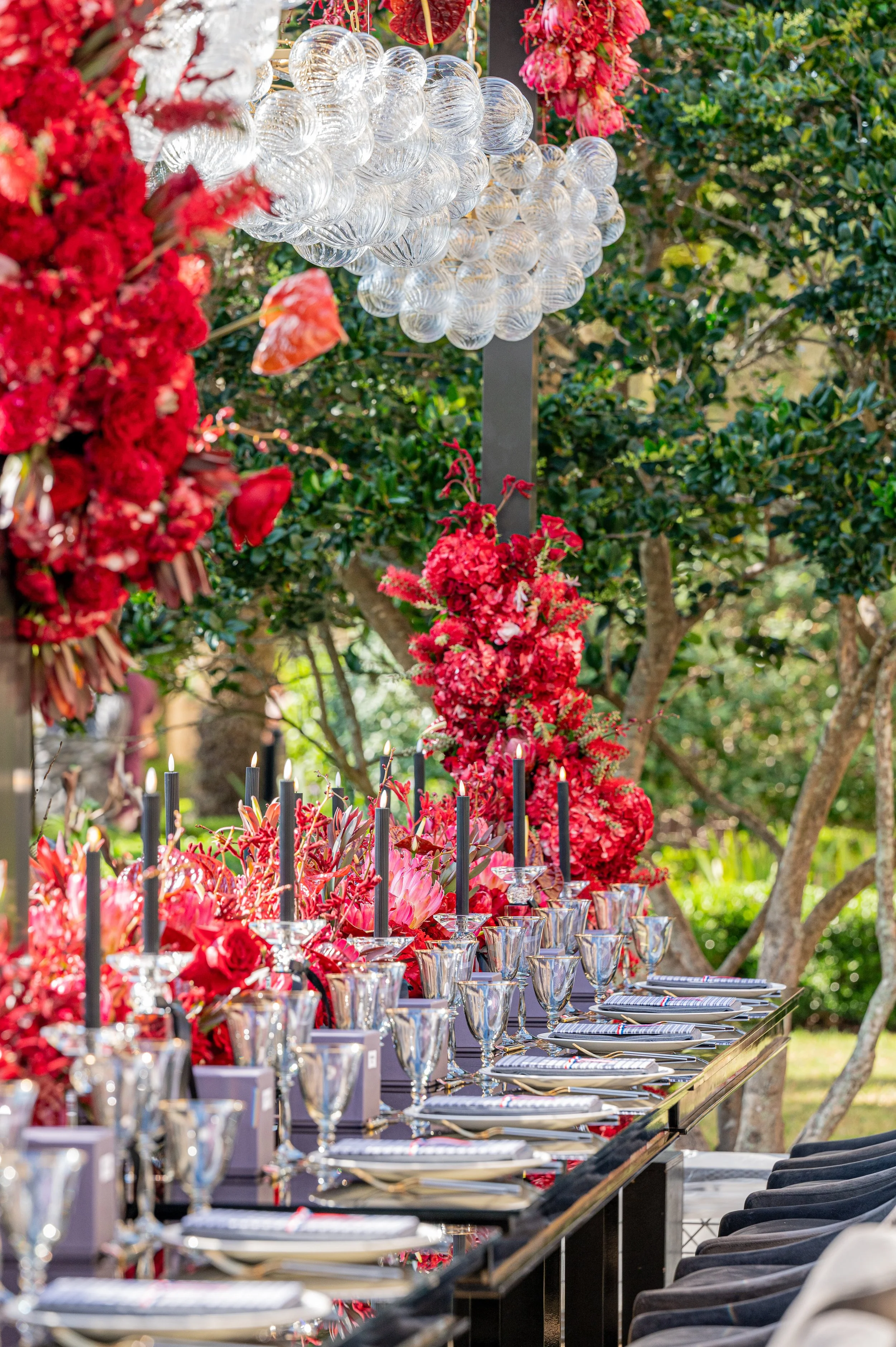 An outdoor dining table decorated with vibrant red flowers, black candles, and silver glassware, set against a lush green garden backdrop with hanging glass orb decorations.