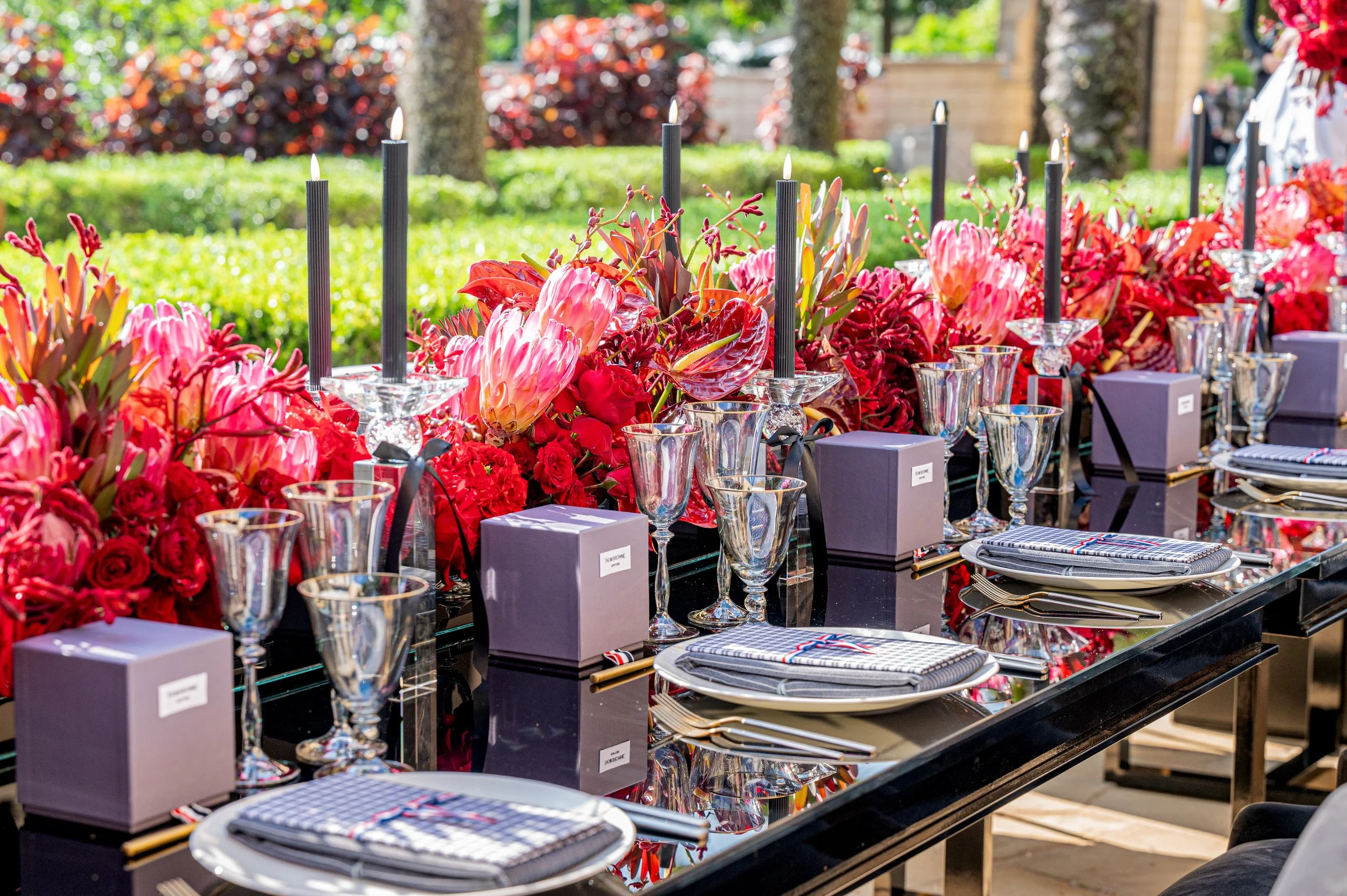 Elegant outdoor dining table set with pink and red flowers, black candles, glassware, and neatly arranged plates and napkins, surrounded by greenery.