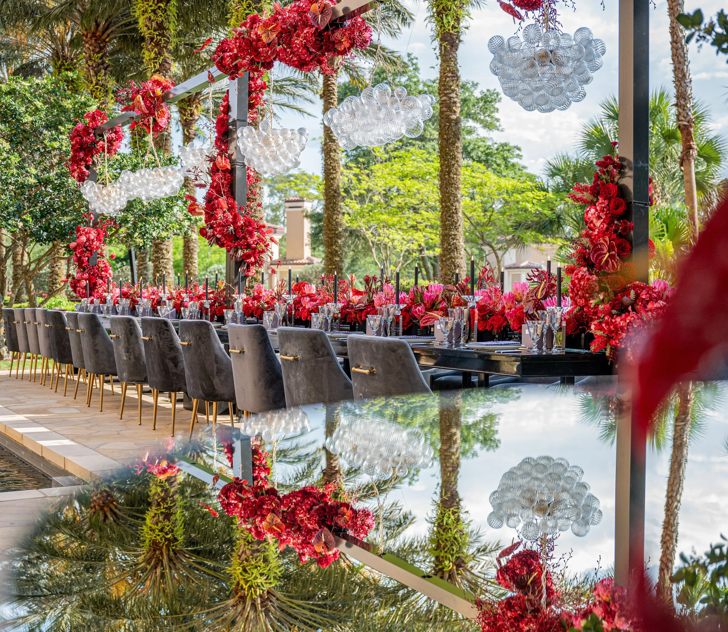 A beautifully decorated outdoor dining table set with pink and red flowers, glassware, and candles, surrounded by lush greenery and palm trees.