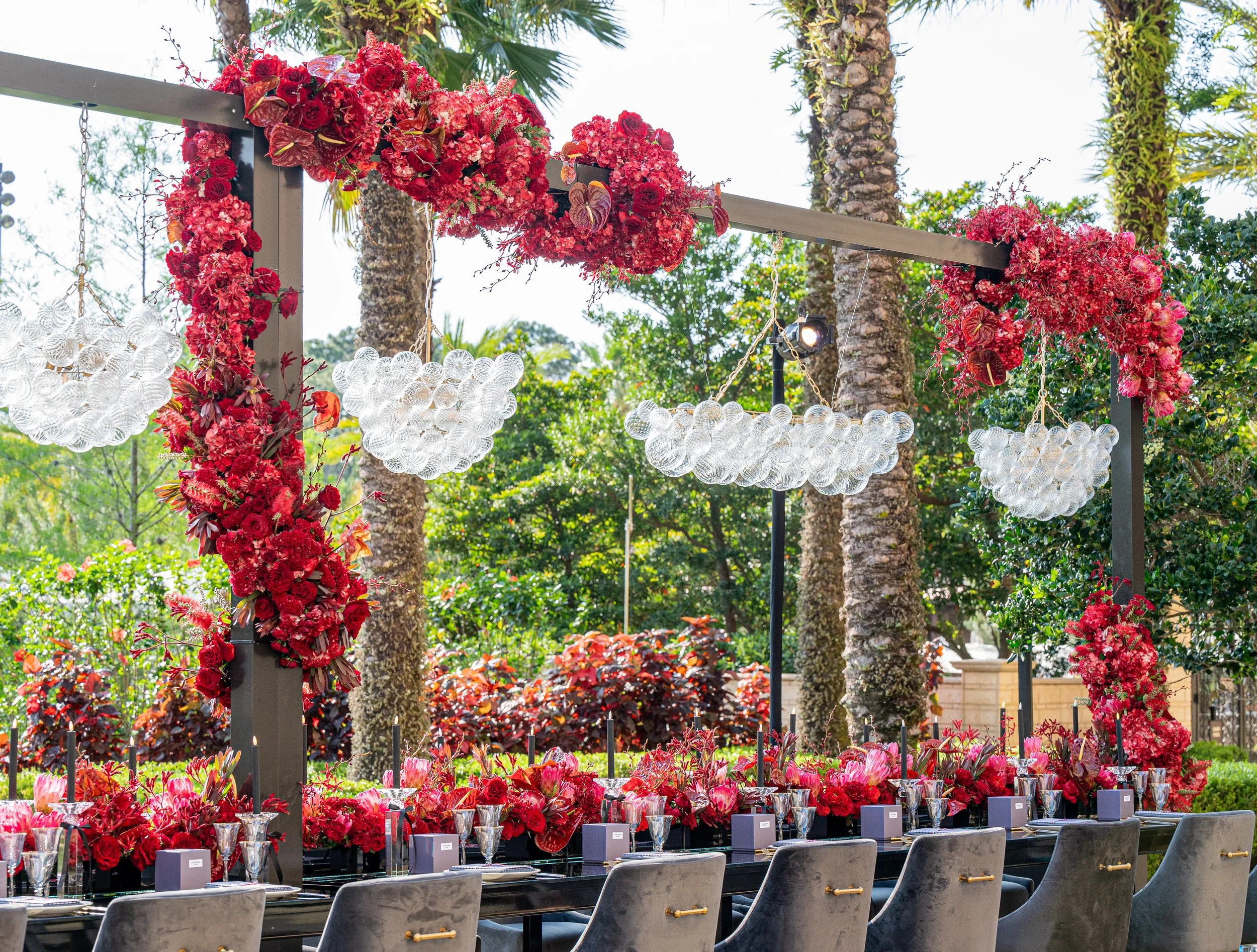 Elegant outdoor dining setup with a long table, decorated with red and pink floral arrangements, crystal glassware, and modern chairs, set against a lush garden with palm trees and greenery.
