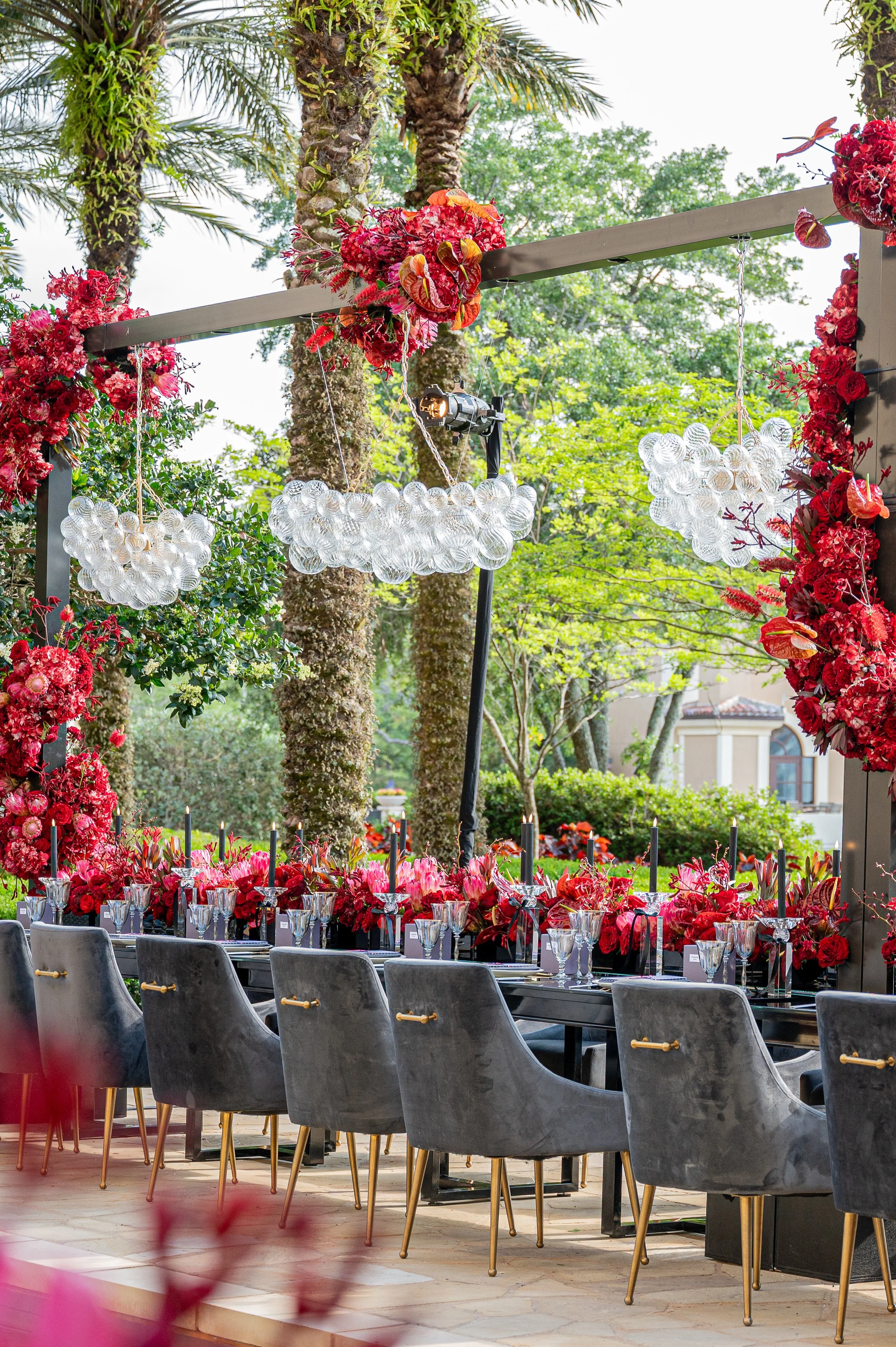 Outdoor dining area decorated with red flowers, black candlesticks, and modern glass chandeliers, set against lush green trees and palm trees.