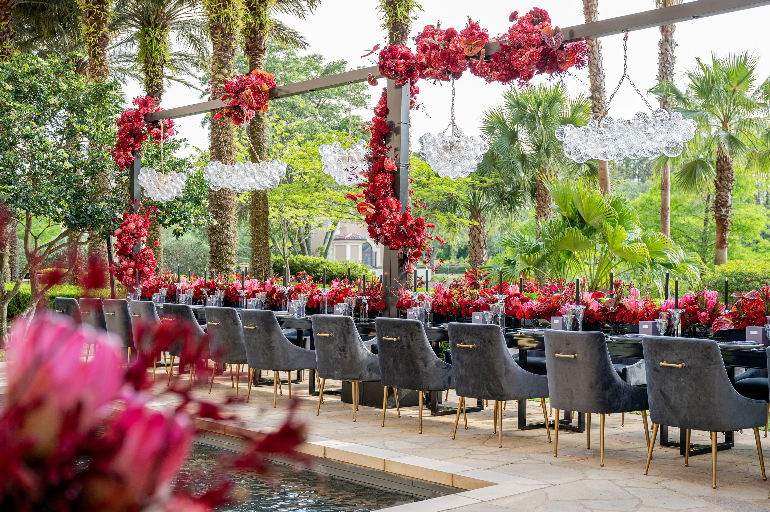 An outdoor dining setup with a long black table, grey velvet chairs, and modern glass cloud-shaped chandeliers, decorated with red flowers and lush green trees in the background.
