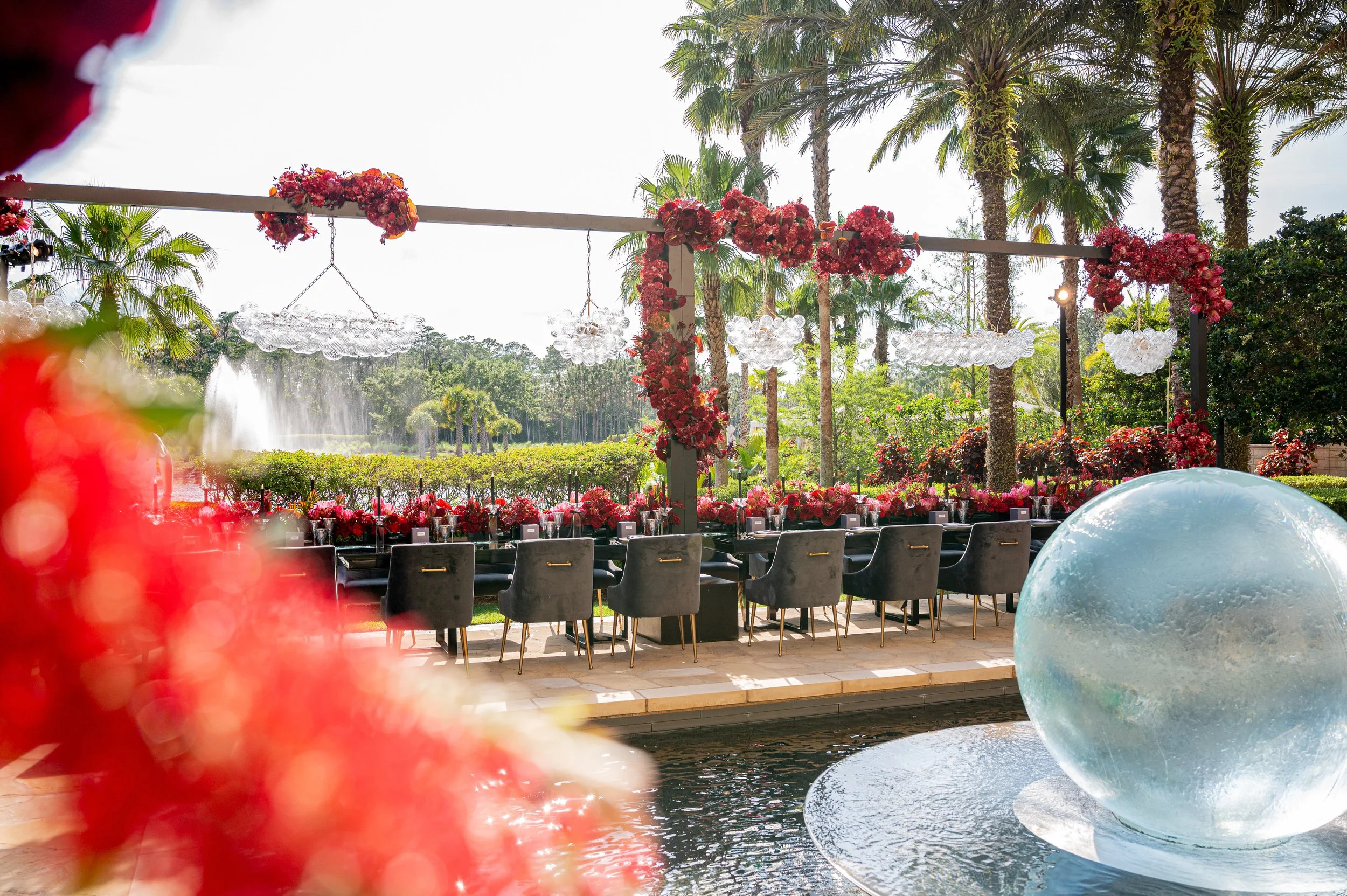 Outdoor event setting with a long table decorated with pink and red floral arrangements, black chairs, and a large water sphere in the foreground. Behind the table, there are tall palm trees, a water fountain, and lush greenery under a partly cloudy 