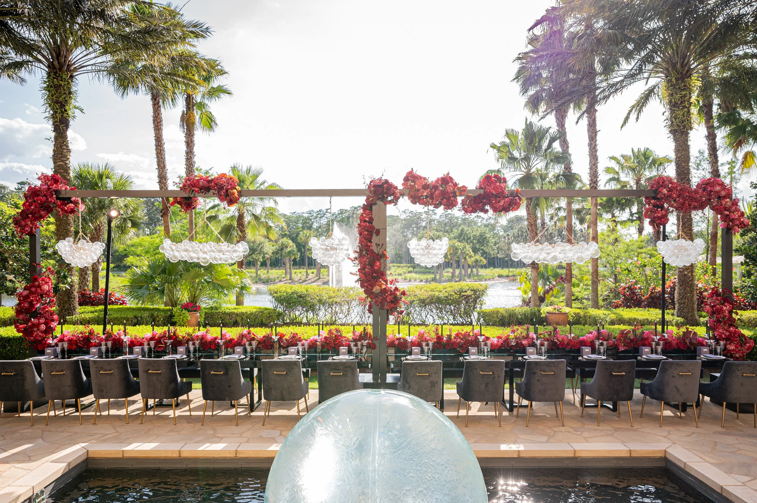 Outdoor wedding or event setup with a long table decorated with pink flowers and surrounded by black chairs, set against a backdrop of palm trees, greenery, and a lake, under a partly cloudy sky.