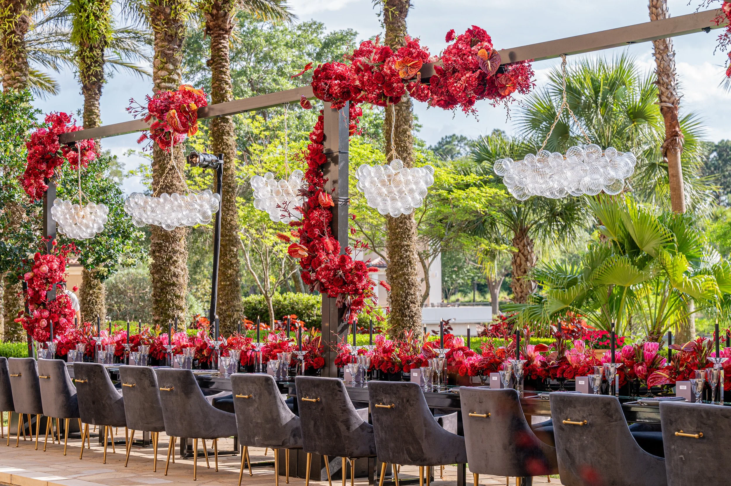 A long outdoor table decorated with red flowers, pink flowers, and greenery, set with glasses and black candles in a lush garden for a celebration or event. Overhead, pink floral arrangements and glass bubble chandeliers are suspended, creating an el