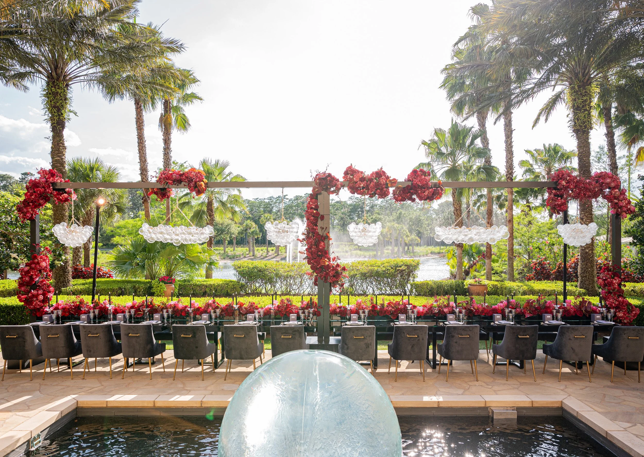 Elegant outdoor dining table set for a special event, decorated with pink and red flowers, arranged in front of a scenic view of tall palm trees, a pond, and lush greenery, with a white fountain in the foreground. Four Seasons Orlando