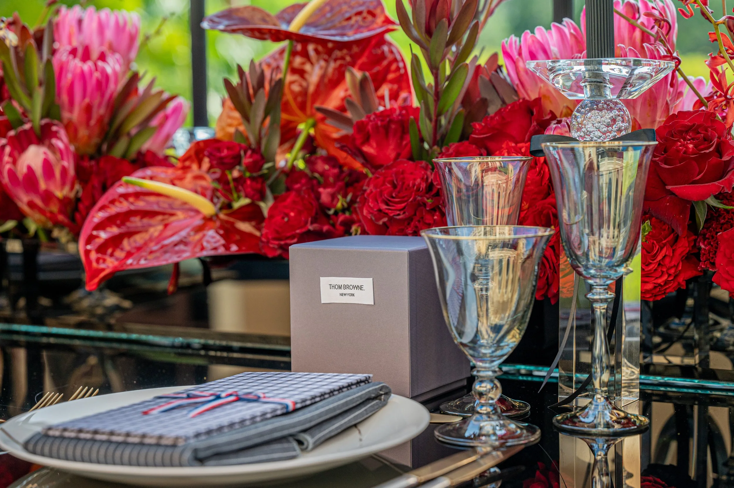 A table setting with a gray box labeled 'Thom Browne New York', three clear glass vases, and a white plate with a folded checkered napkin. The background features a vibrant arrangement of red and pink flowers.