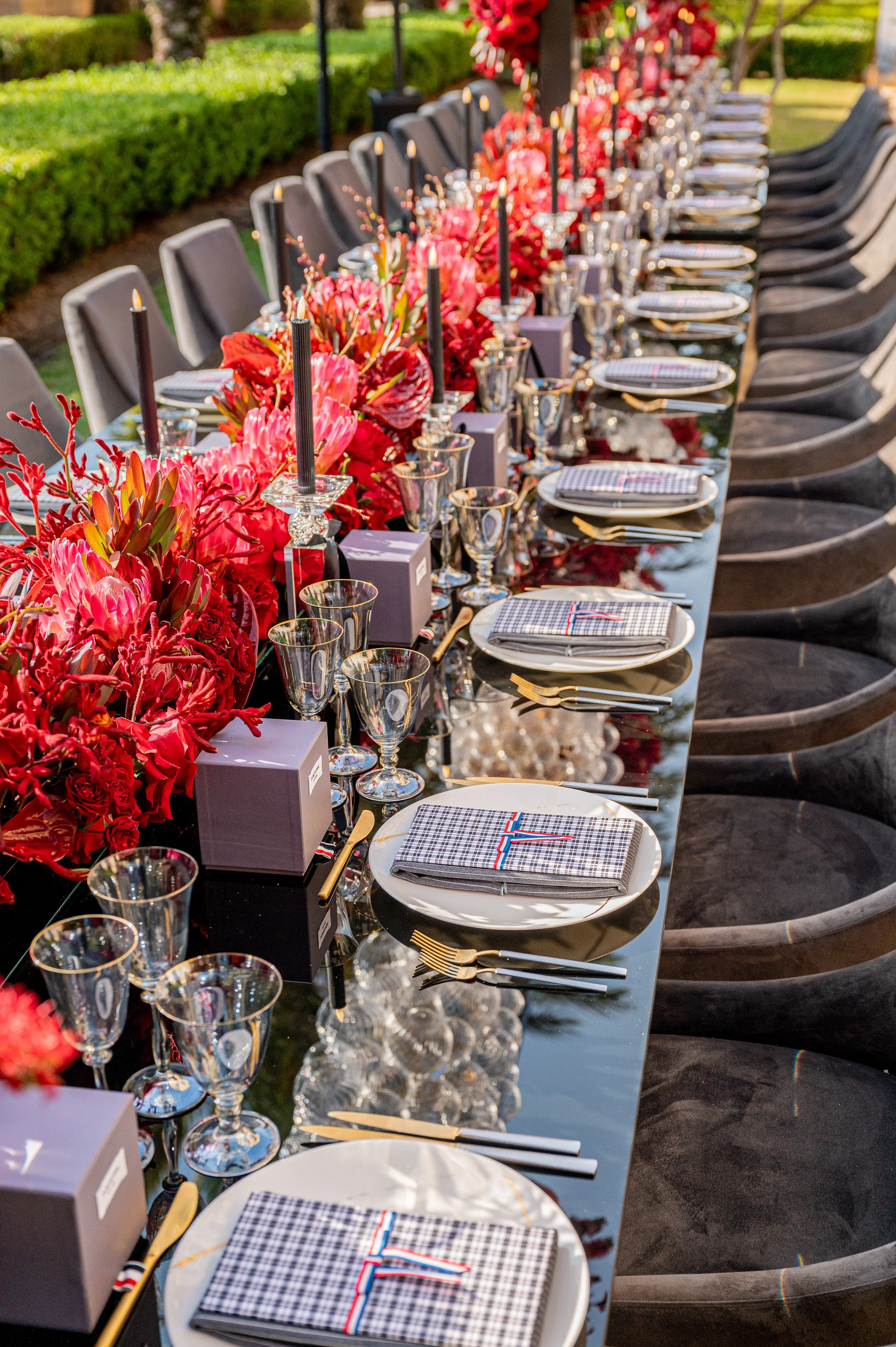 A long outdoor dining table decorated with pink and red floral arrangements, black candles, glassware, and neatly folded napkins, set for a formal event or celebration.