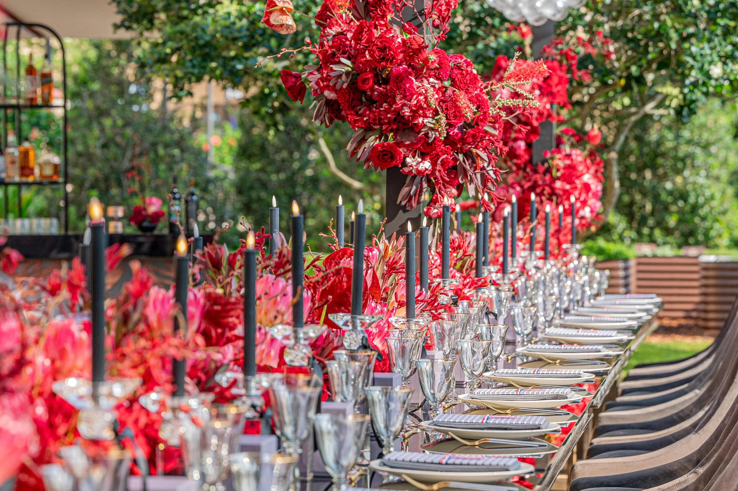 A long outdoor table set for a formal event, decorated with elaborate red floral centerpieces and lit black candles, with place settings including plates, glasses, and cutlery, surrounded by chairs and lush greenery.