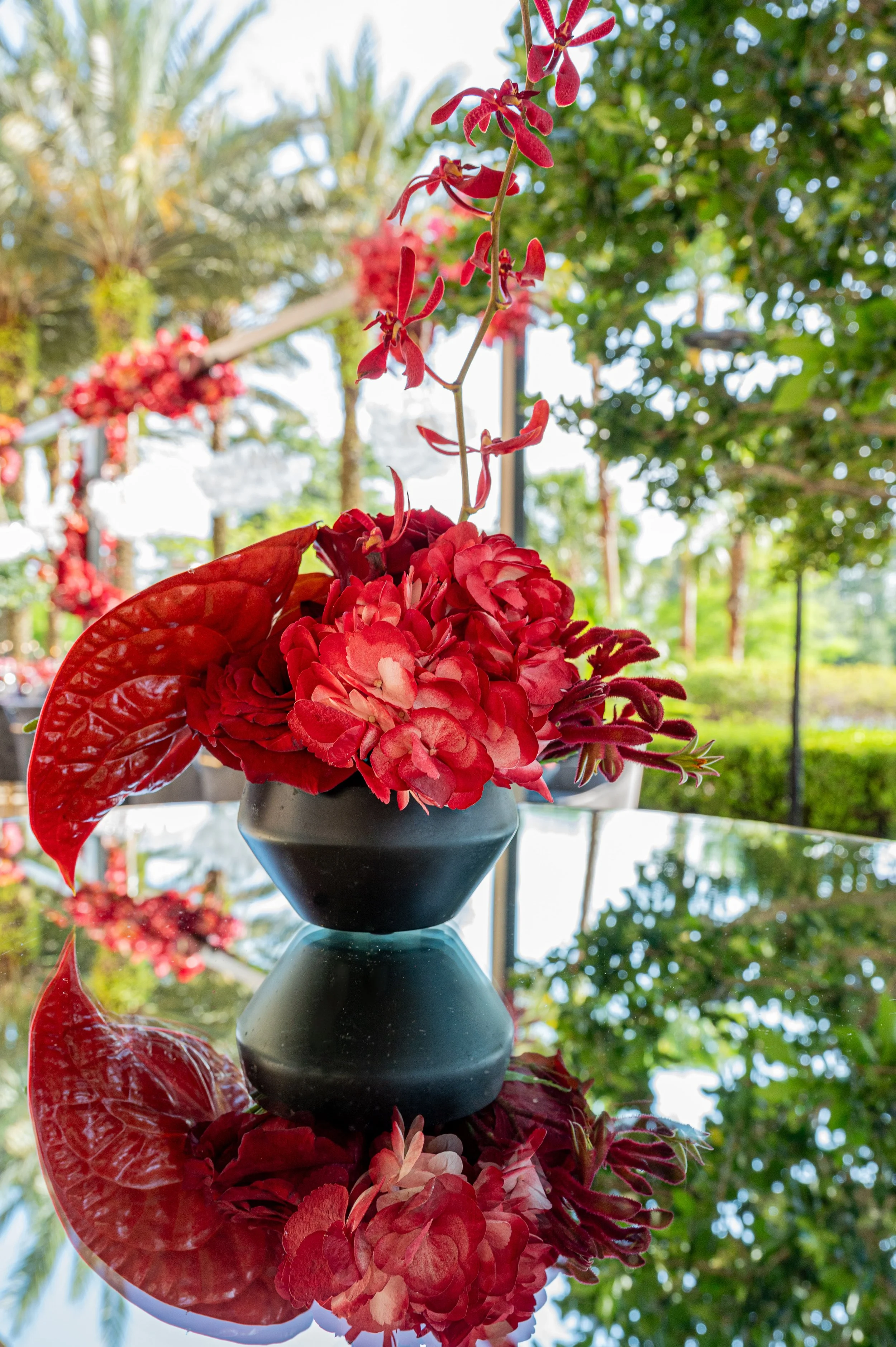 A black flower vase with red artificial flowers, including hydrangeas and anthuriums, sitting on a reflective glass surface outdoors with lush green trees in the background.