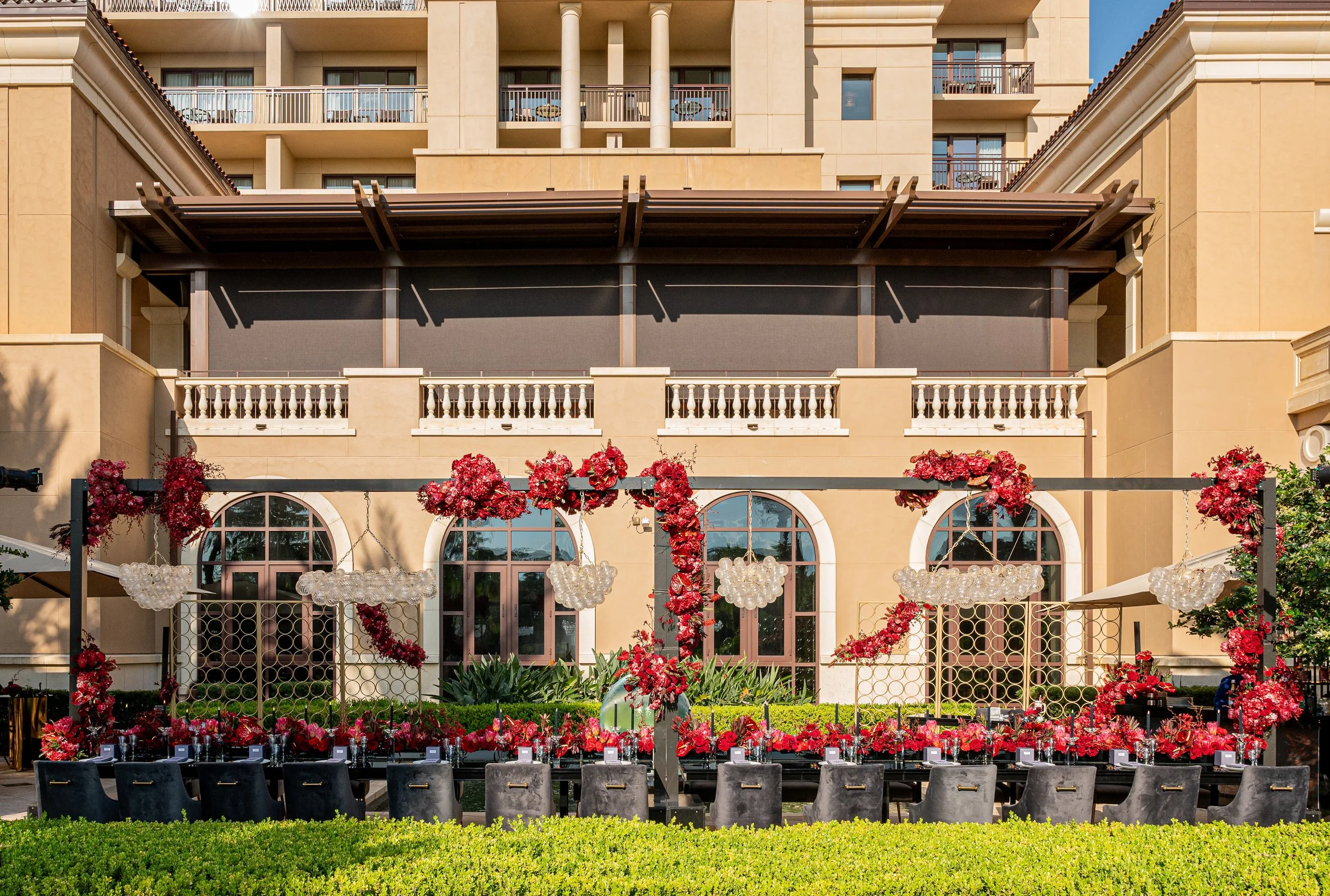 Outdoor wedding or event setup with long tables, black chairs, and red floral decorations, ornate chandeliers, and a beige multi-story building in the background. Four Seasons Orlando