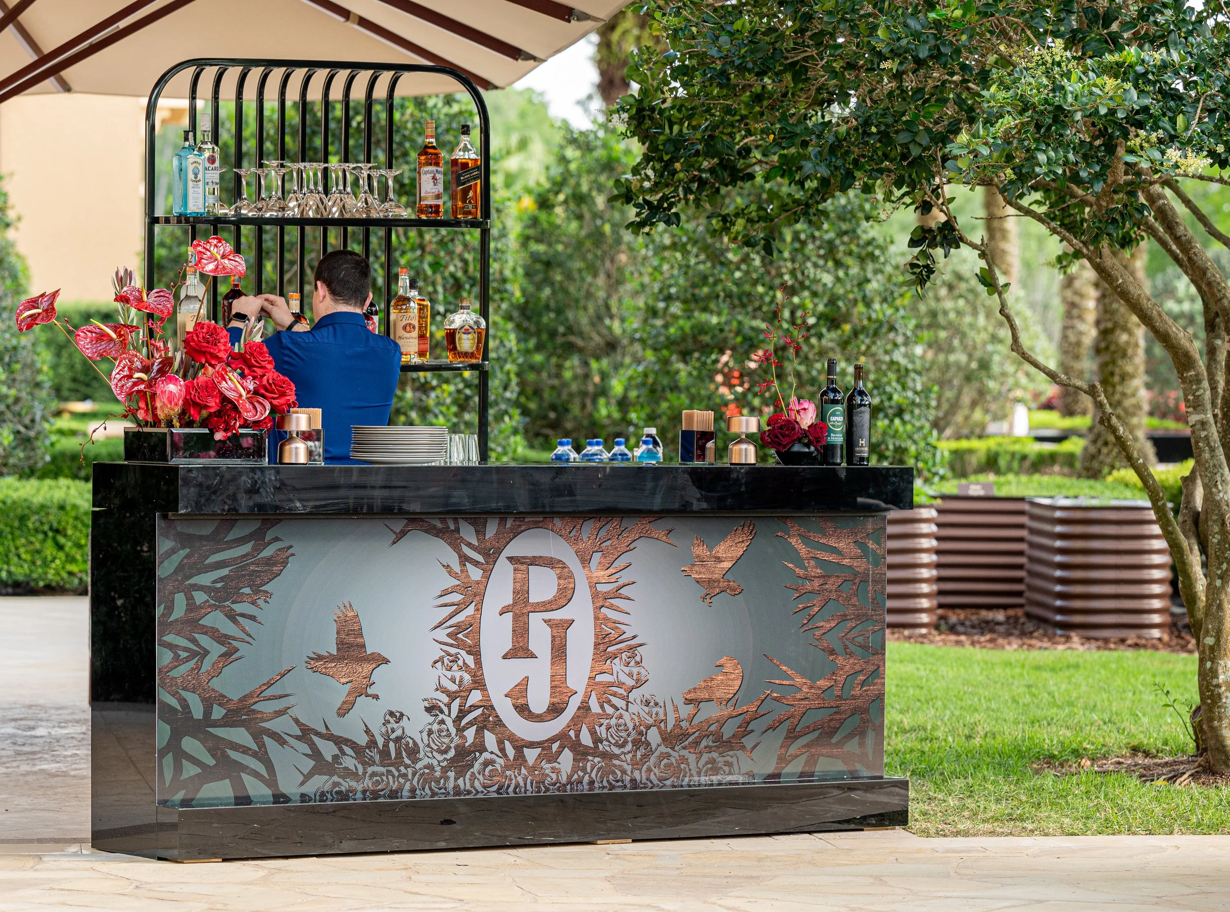 An outdoor bar with a black marble top and decorative front panel displaying the initials "PJ" surrounded by birds and floral motifs. The bar has a person in a blue shirt behind it, preparing drinks. There are bottles of alcohol on shelves above the 