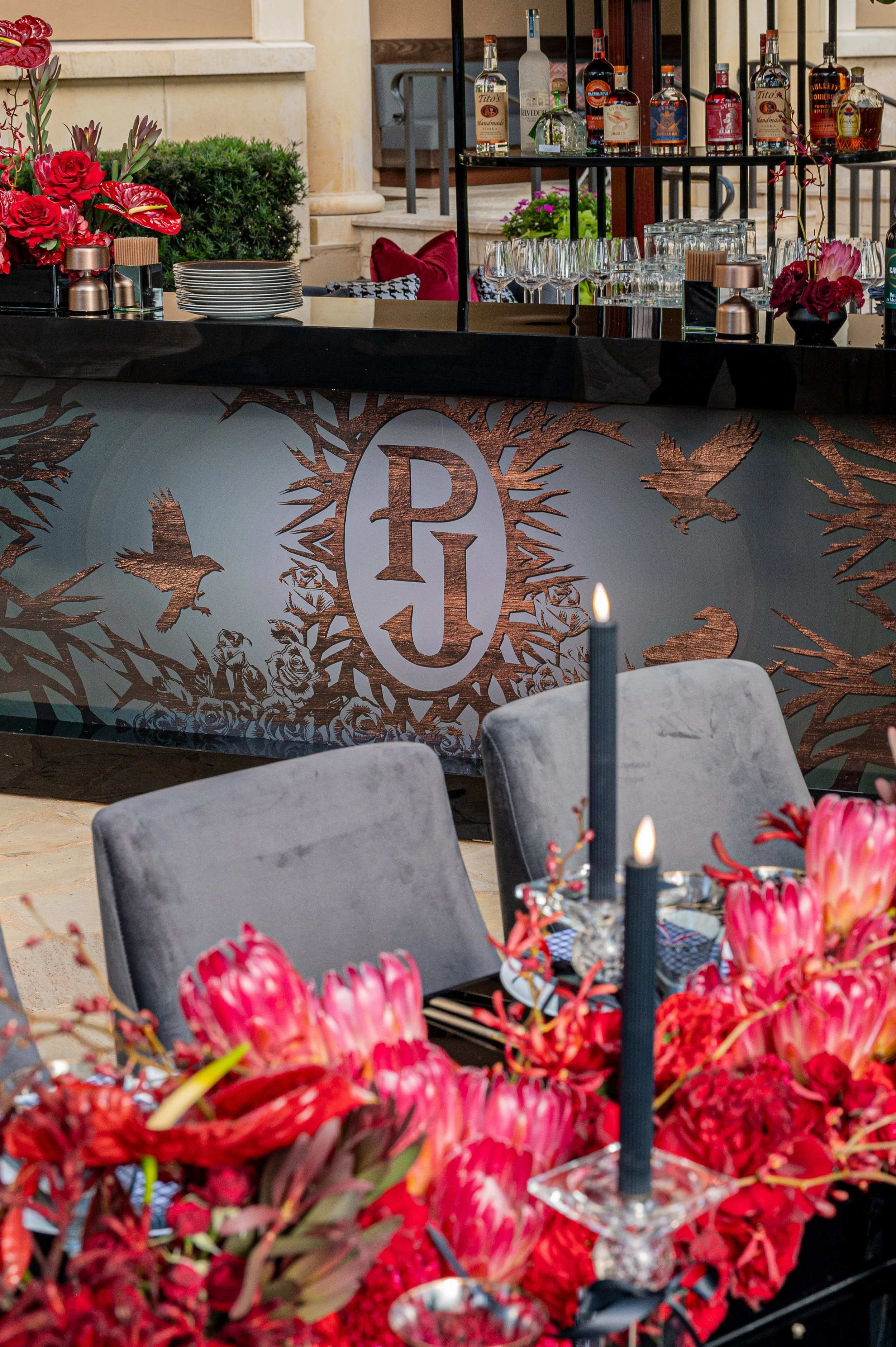 A bar with a black bar top and a decorative banner with a logo surrounded by birds and flowers, set with glasses and bottles behind it. In the foreground, a table with pink flowers and black candles.