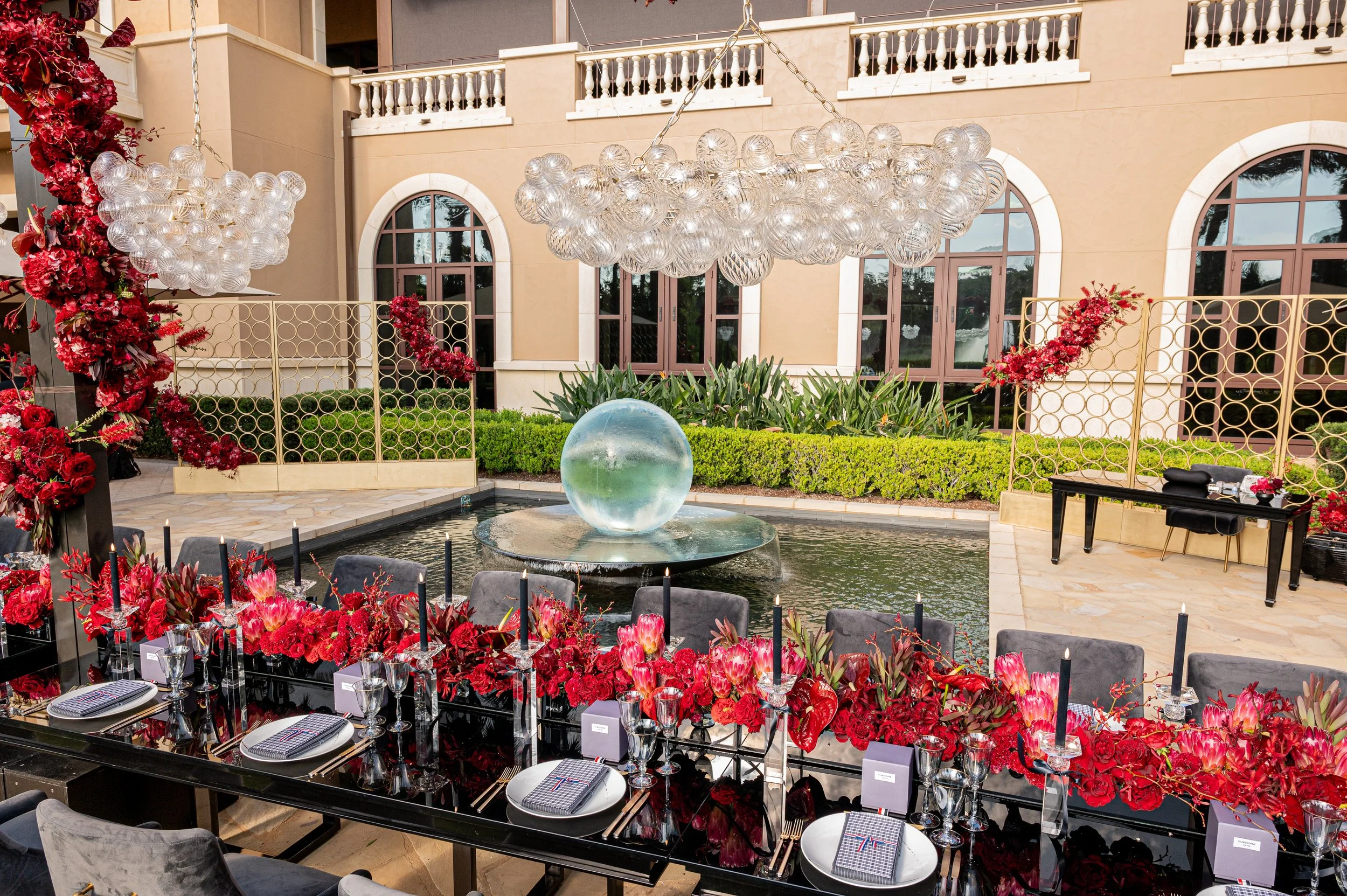 Elegant outdoor event setup with a long table decorated with red flowers and black candles, overlooking a water fountain with a glass sphere, in front of a beige building with arched windows and a balcony. Four Seasons Orlando