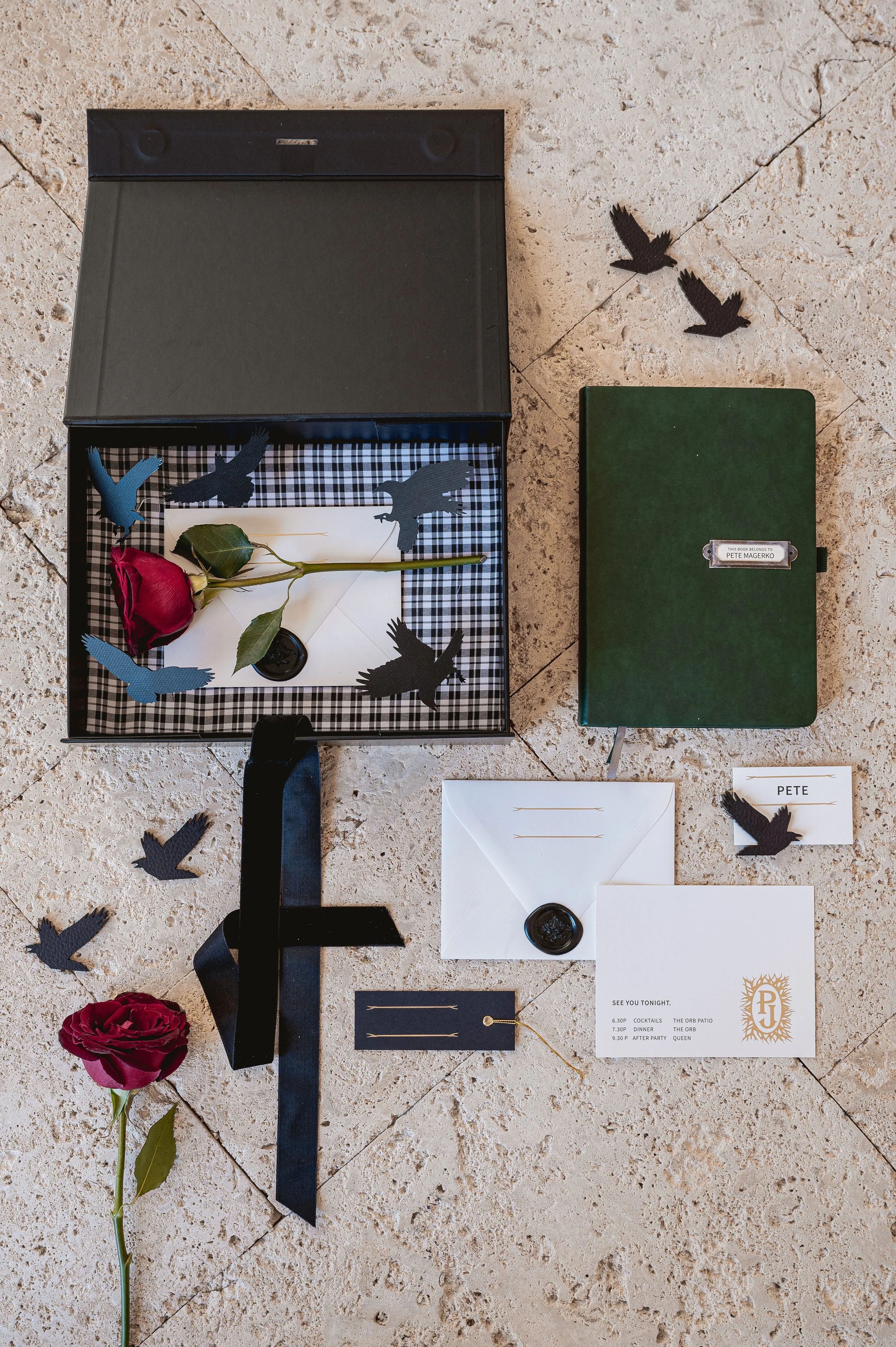 A black gift box with a plaid lining, containing a red rose, white envelope, and cutout black and blue bird figures, with additional bird cutouts, a green book, and wedding invitation items on a tiled floor.