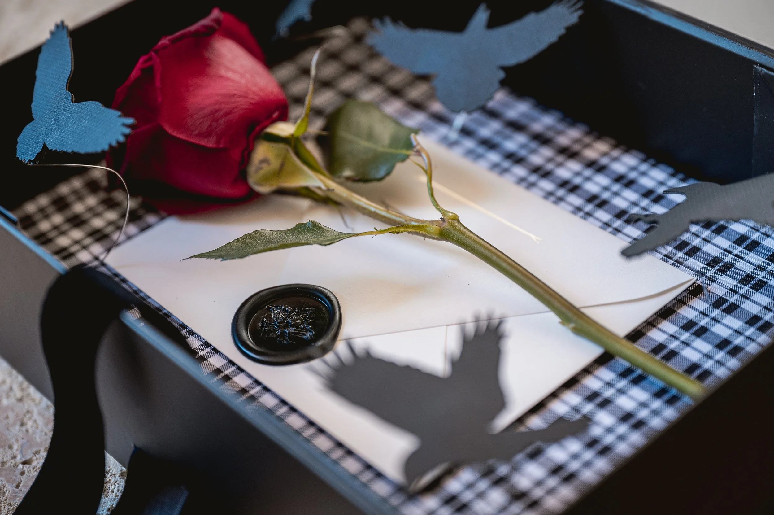 A red rose with green leaves placed on a white card inside a black gift box, decorated with blue paper butterflies and black and white checkered patterned paper, with a black wax seal nearby.