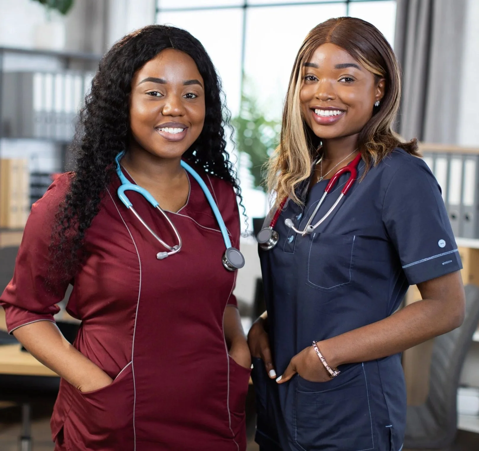 Two smiling female healthcare professionals wearing scrubs and stethoscopes in a medical office setting.