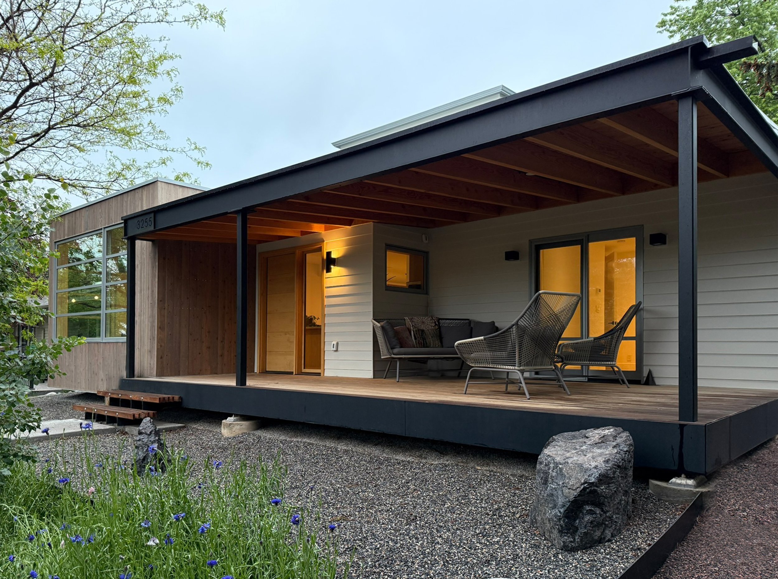 Modern house with a covered wooden deck, outdoor seating, and a landscaped yard with purple flowers.