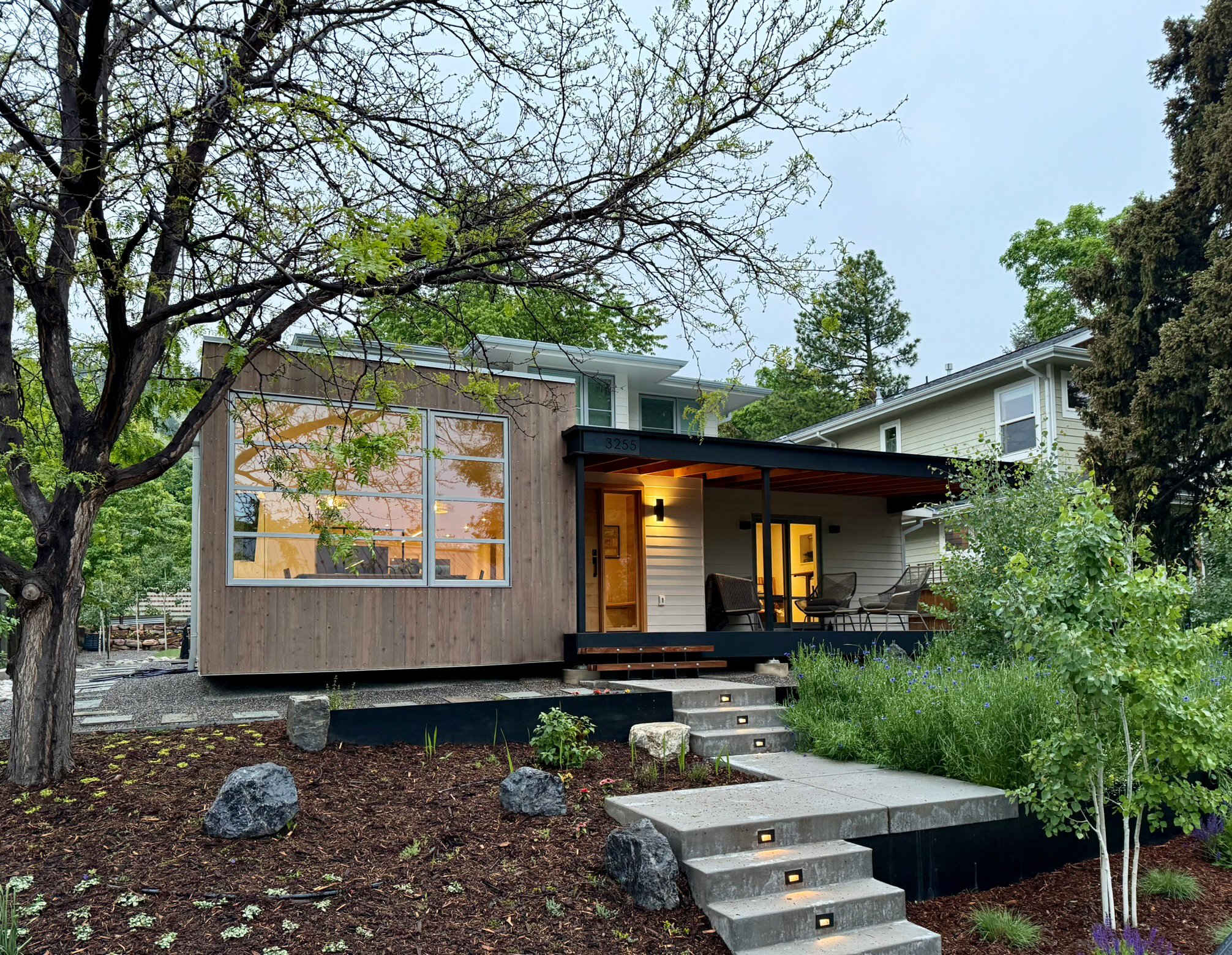 Modern house with large front windows, porch with outdoor furniture, surrounded by trees and landscaped garden with rocks and mulch.